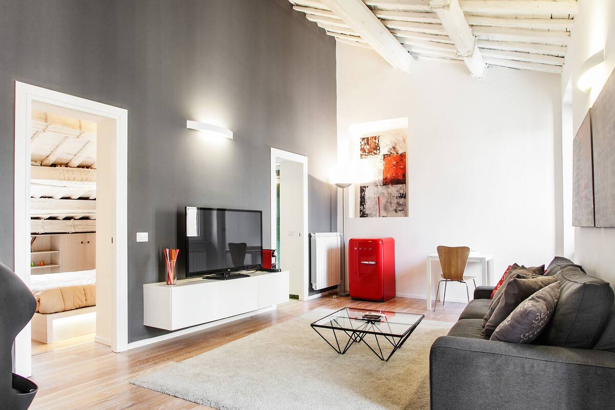 A modern living room with a flat-screen TV, a red refrigerator, a glass coffee table, and a grey sofa.