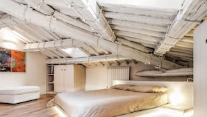 A bedroom with a sloped ceiling, wooden beams, a bed with a beige blanket, a white sofa, and a radiator. - At Home Spanish Steps (Rome)