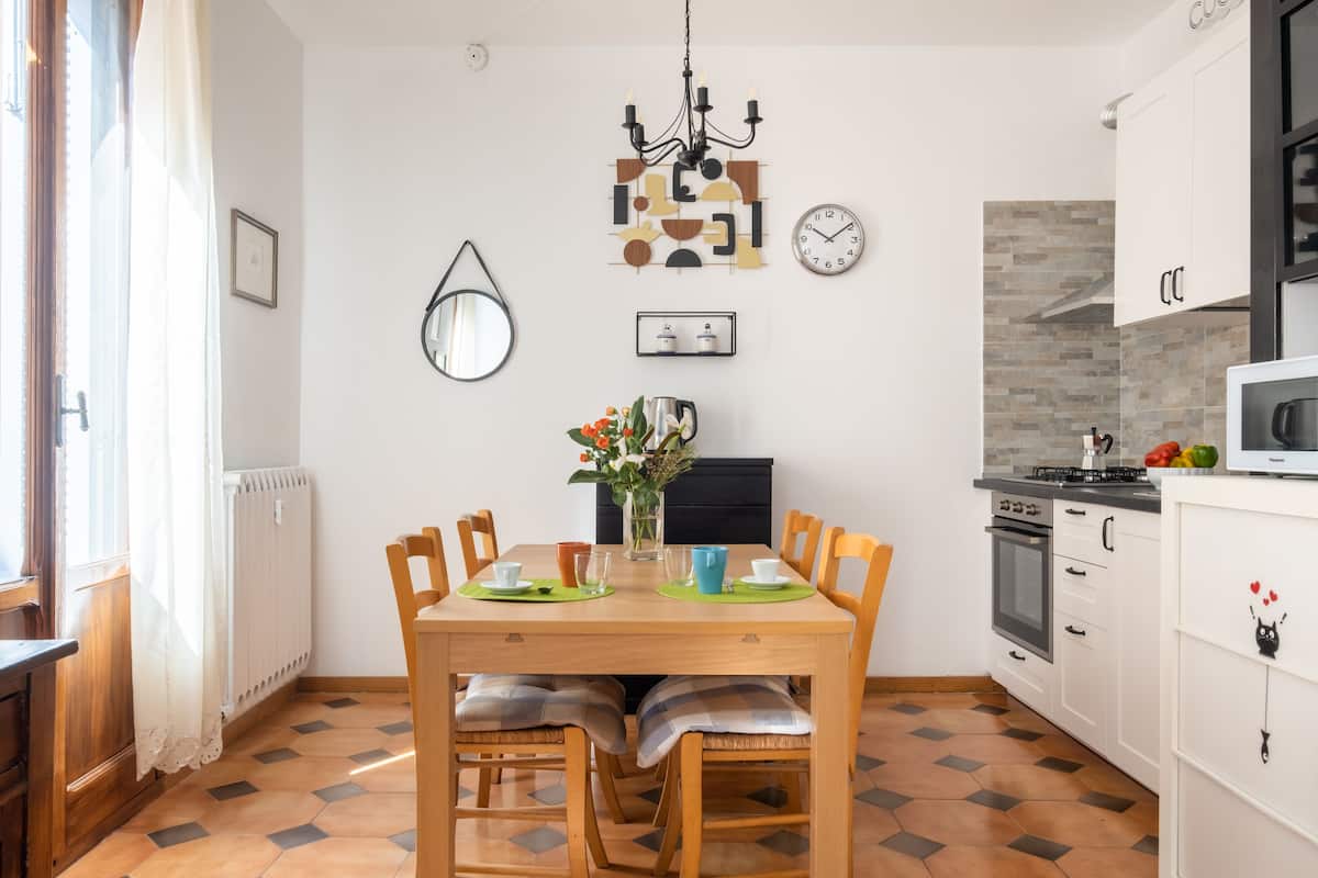 Un coin repas comprenant une table en bois, des chaises, un lustre, une horloge, ainsi qu’une cuisine avec des meubles blancs et un four à micro-ondes.