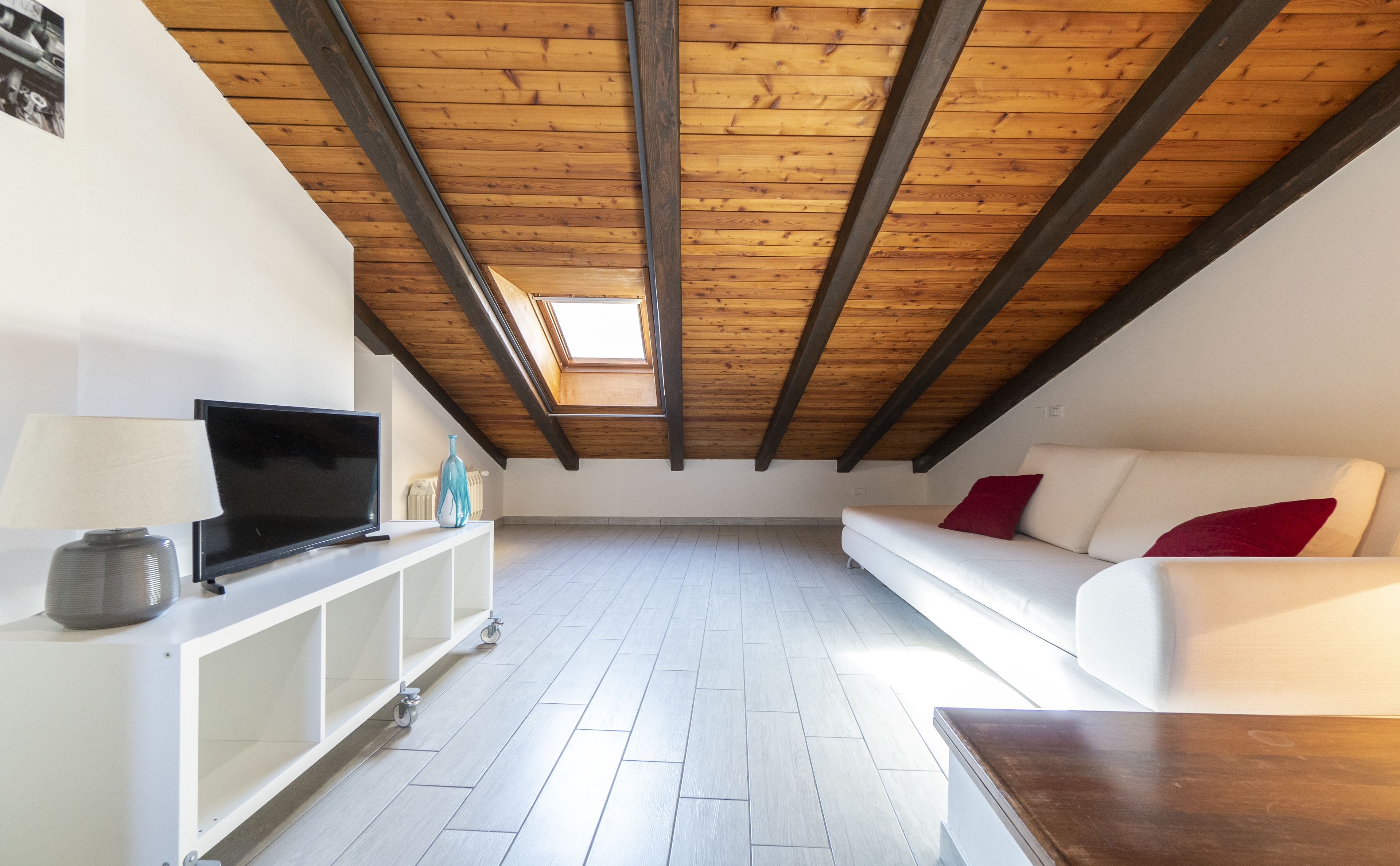 Un ático espacioso con techo de madera, un sofá blanco con cojines rojos, un televisor sobre un mueble blanco y una mesita pequeña.