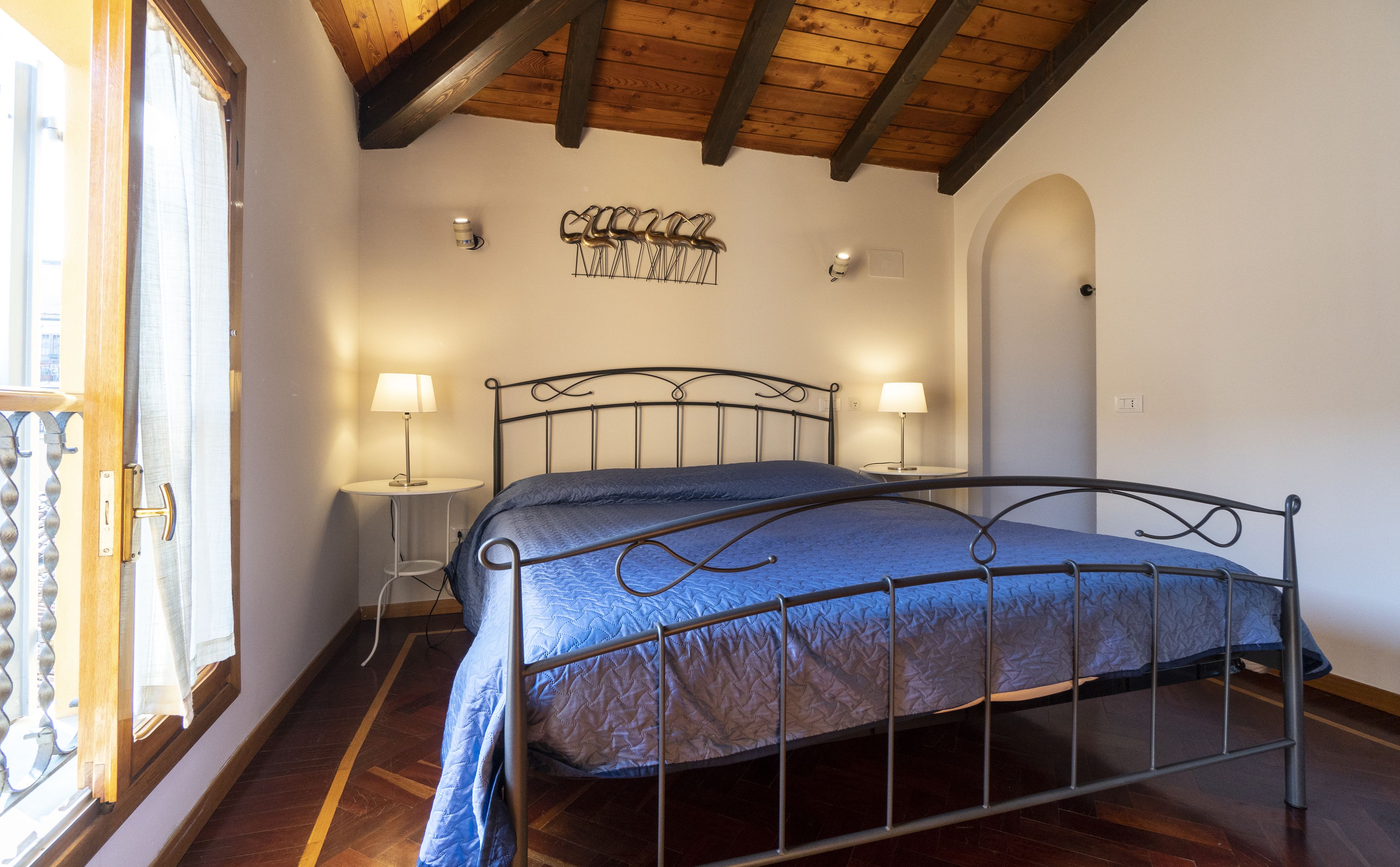 Een slaapkamer met een metalen bedframe, twee bedlampen, een wandlamp en een houten plafond.