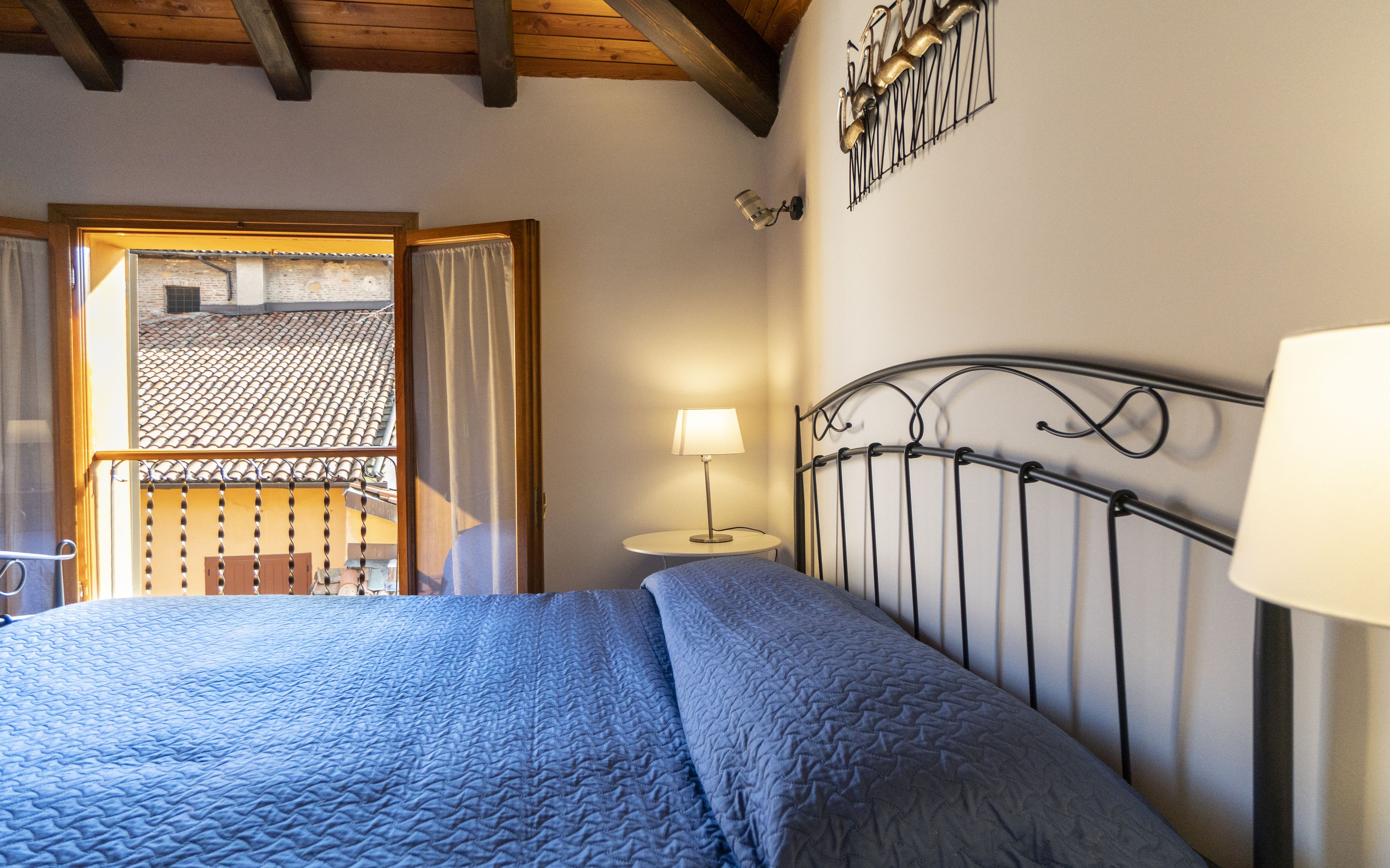 Un dormitorio con cama, mesita de noche con lámpara, ventana con cortinas y vista a un tejado de tejas.