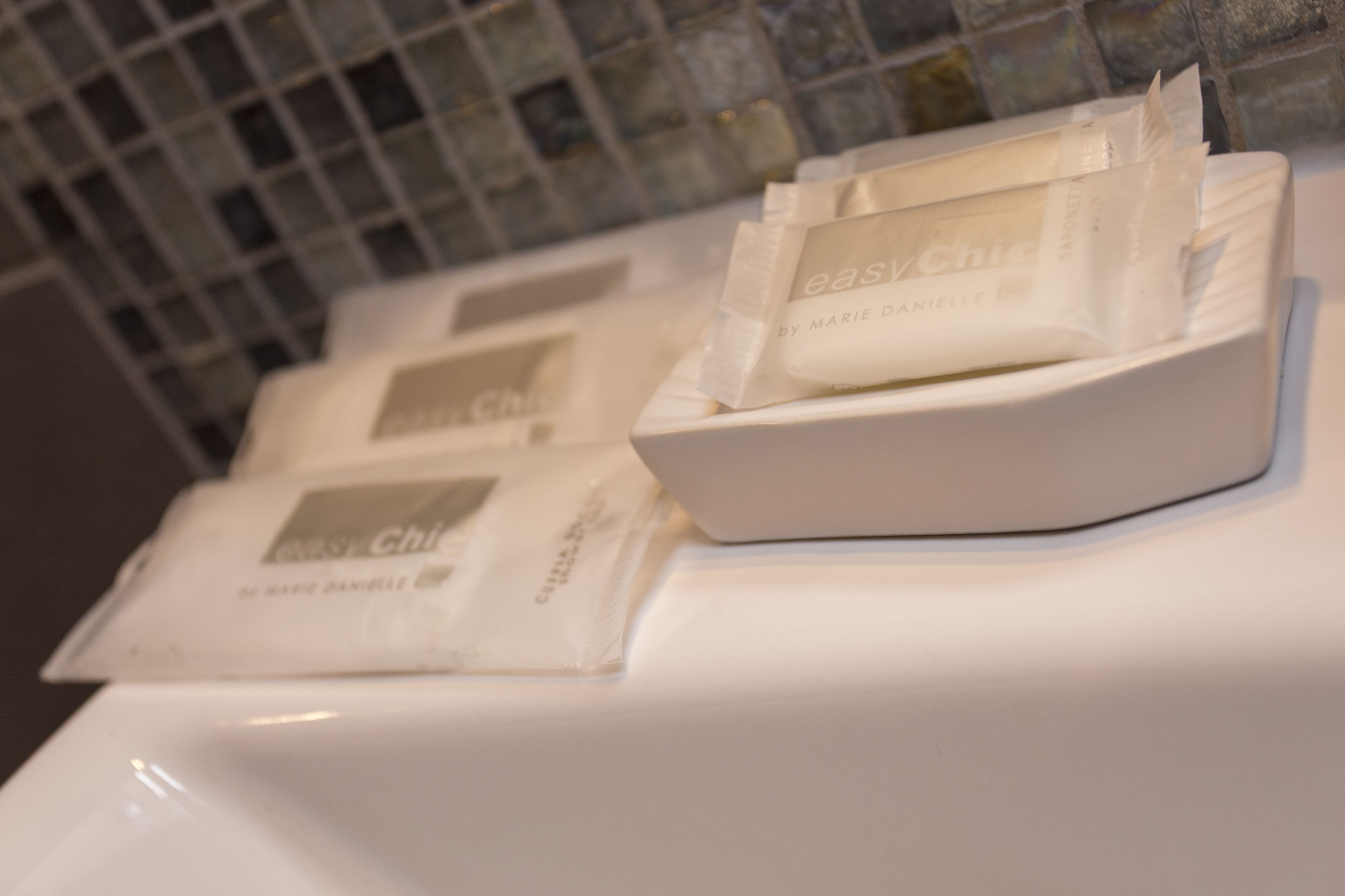 A bathroom with a white sink and a tiled wall. There are two packages of Marie Daniele soap on the sink.