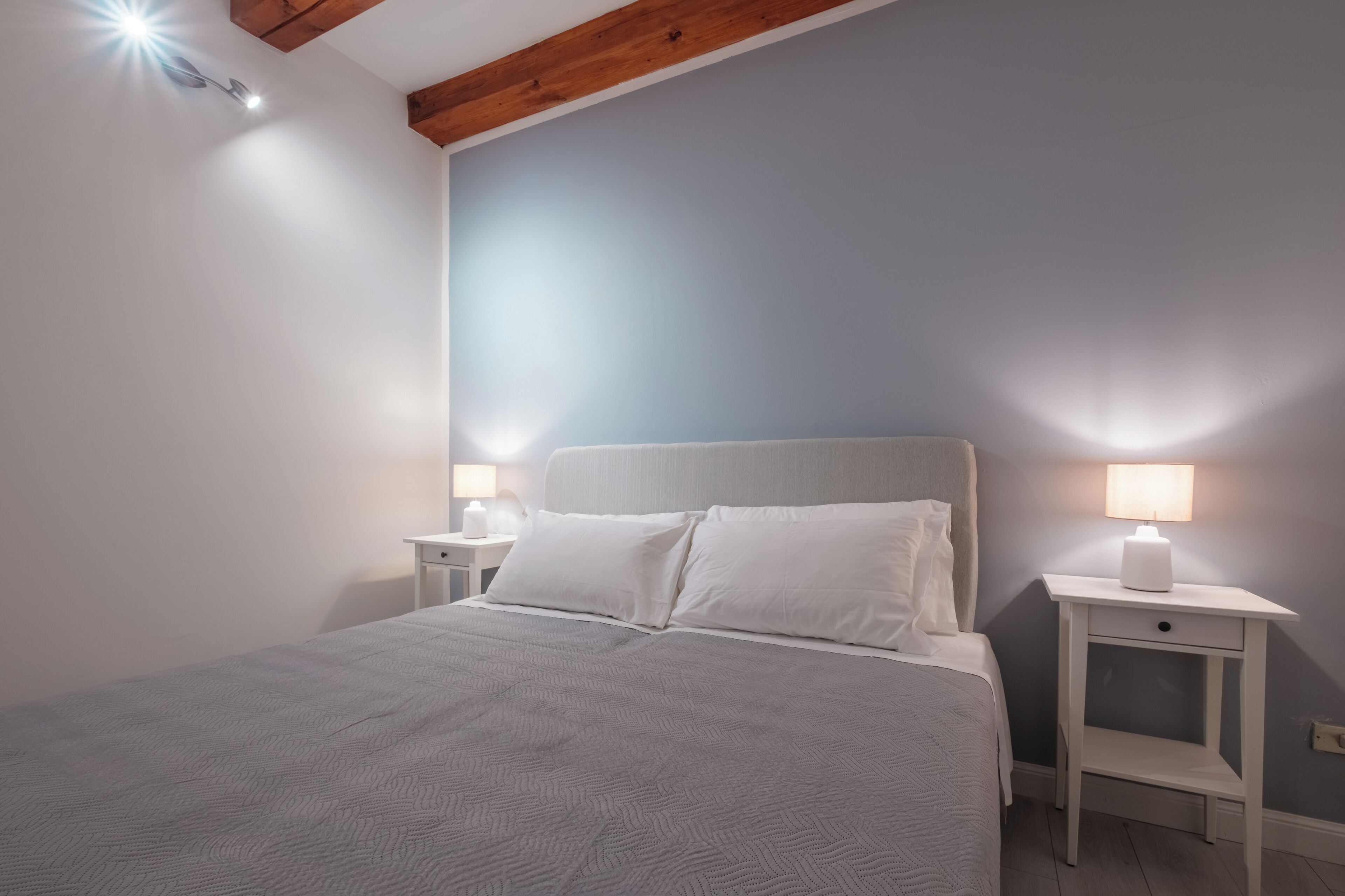 Una cama bien tendida con sábanas blancas, dos mesitas de noche con lámparas y una viga de madera en el techo.
