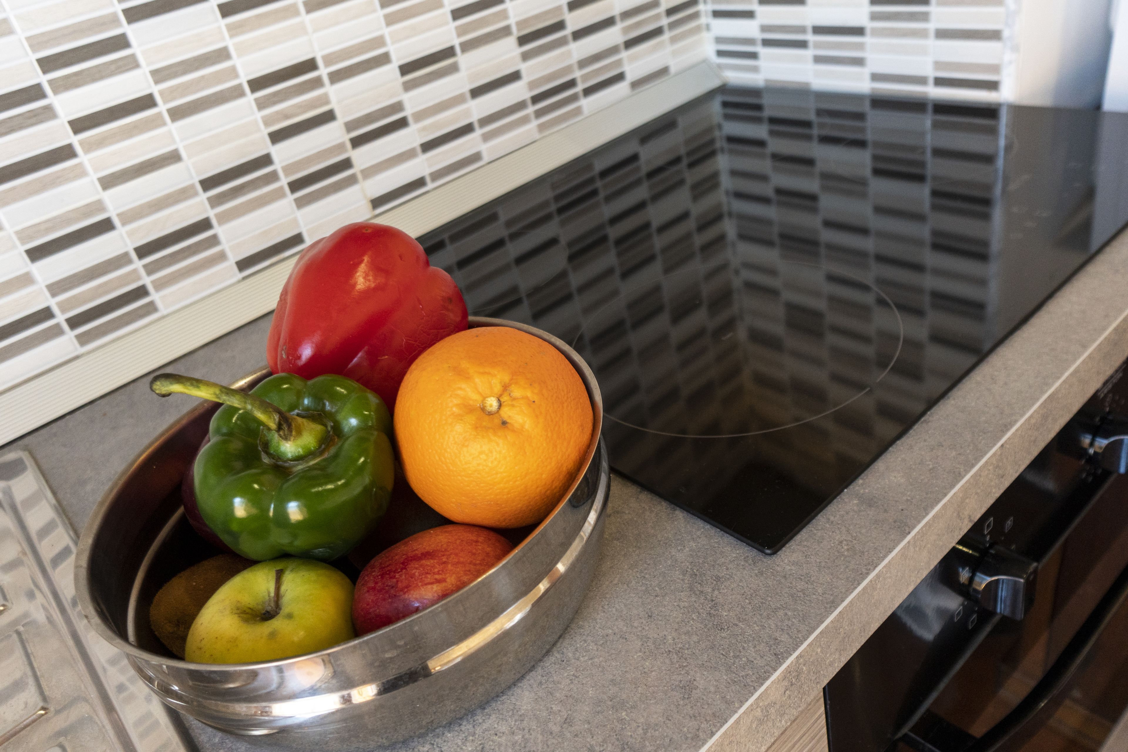 A stainless steel bowl containing a green bell pepper, an orange, a red apple, and a yellow apple on a kitchen countertop.