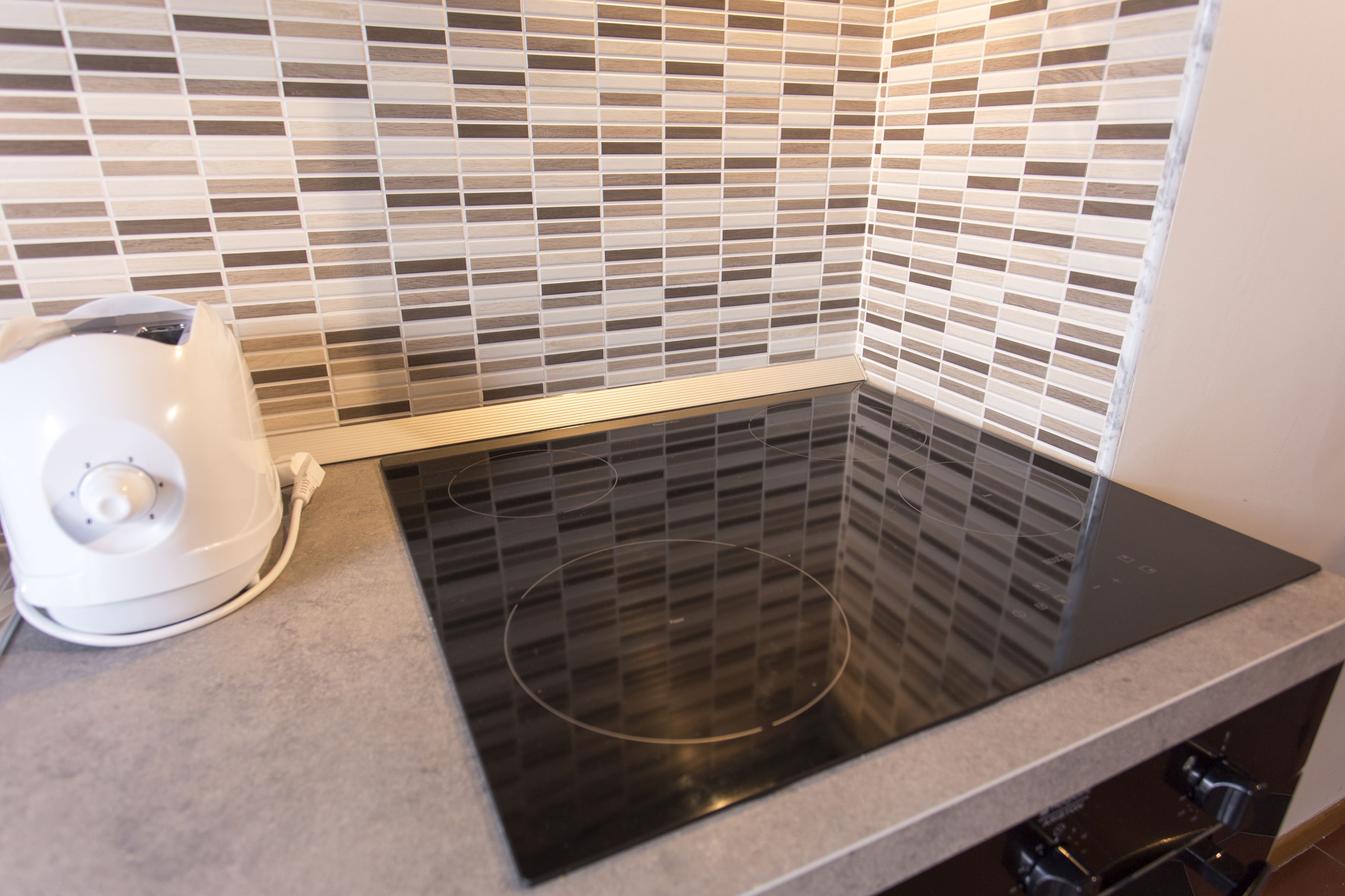 Una cocina con tope de inducción negro, hervidor blanco y salpicadero de azulejos.