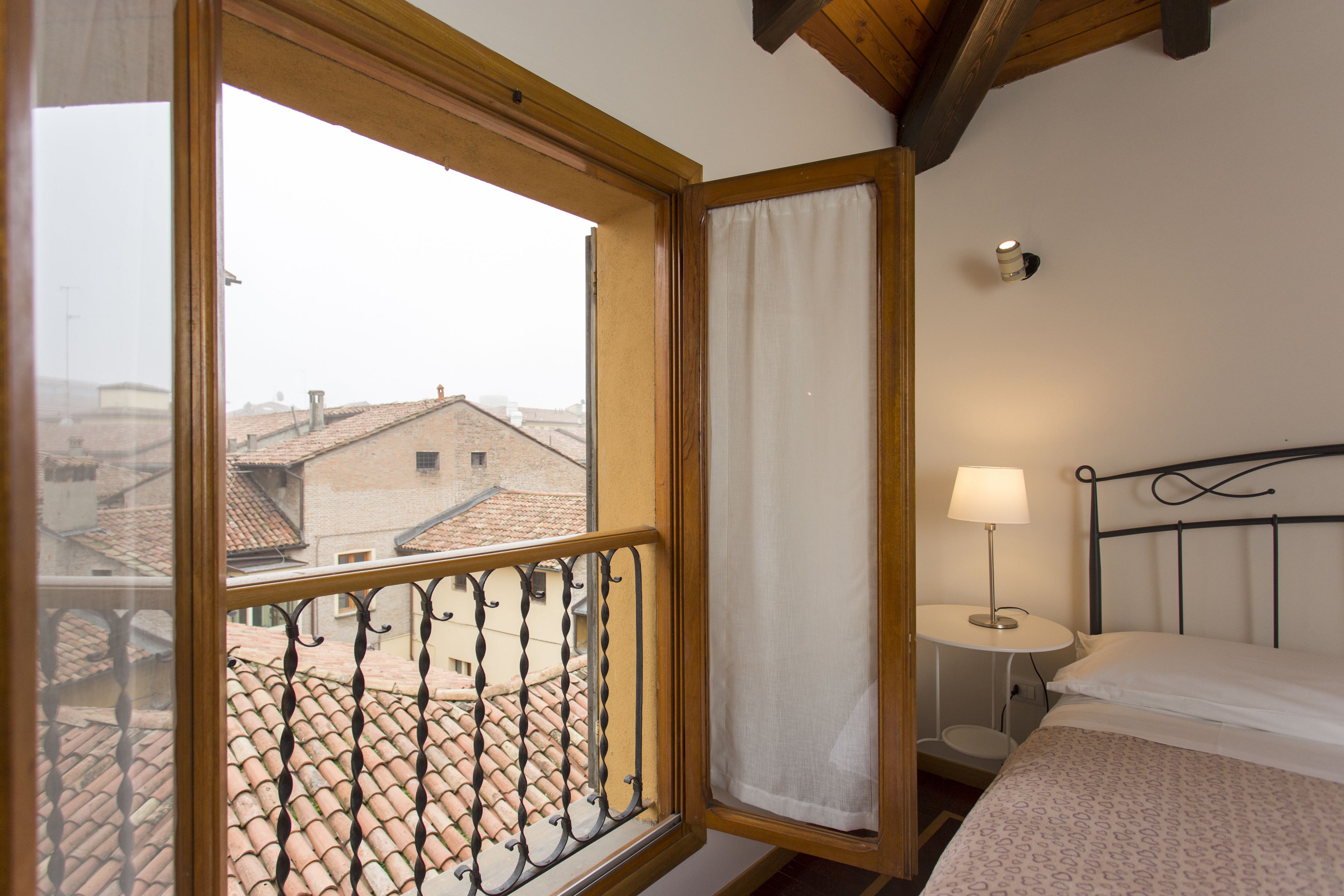 A room with a bed, a bedside table with a lamp, a window with a view of buildings, and a curtain.