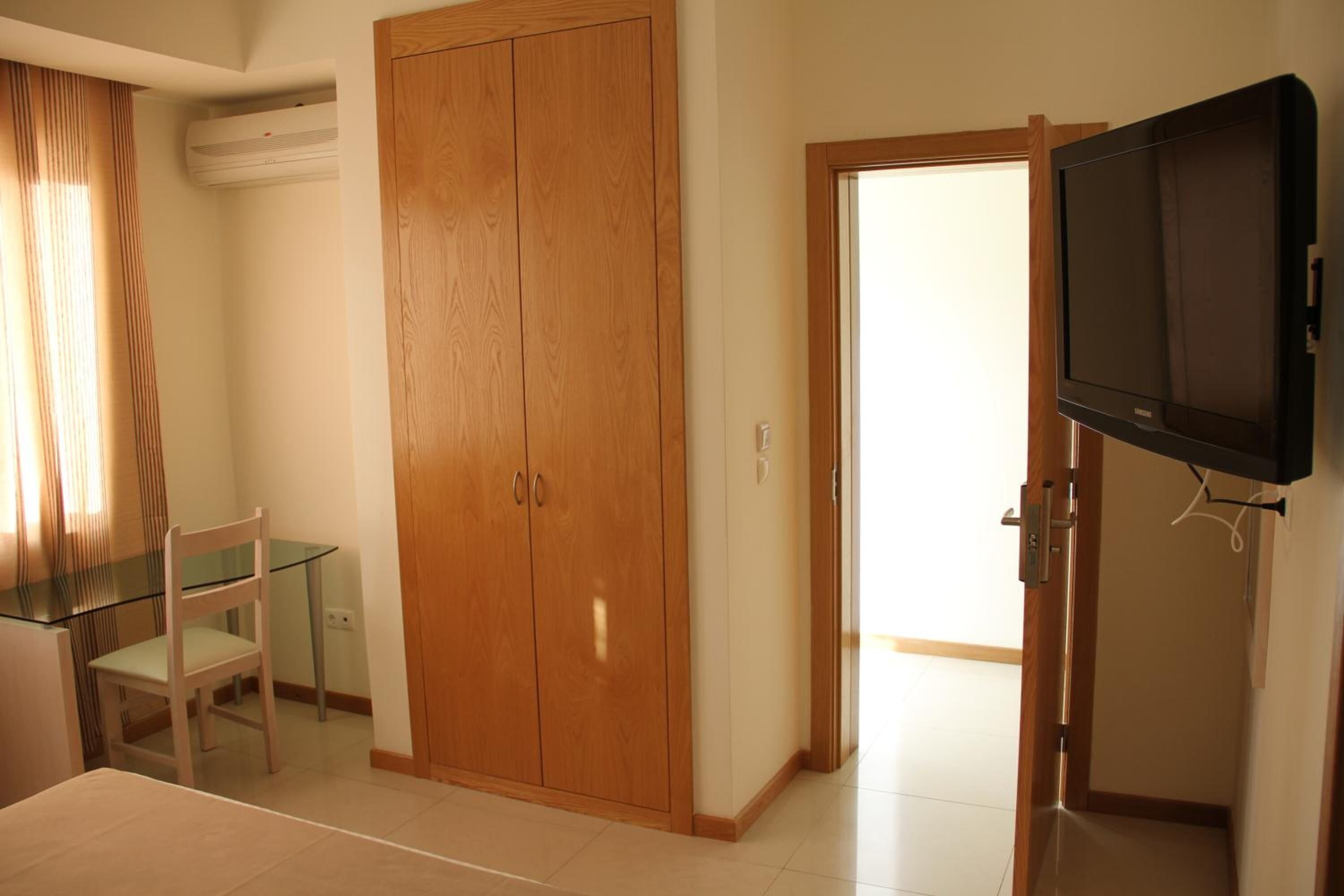 A bedroom with a wooden wardrobe, a glass table with a chair, and a television mounted on the wall.