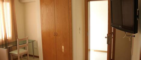 A bedroom with a wooden wardrobe, a glass table with a chair, and a television mounted on the wall.
