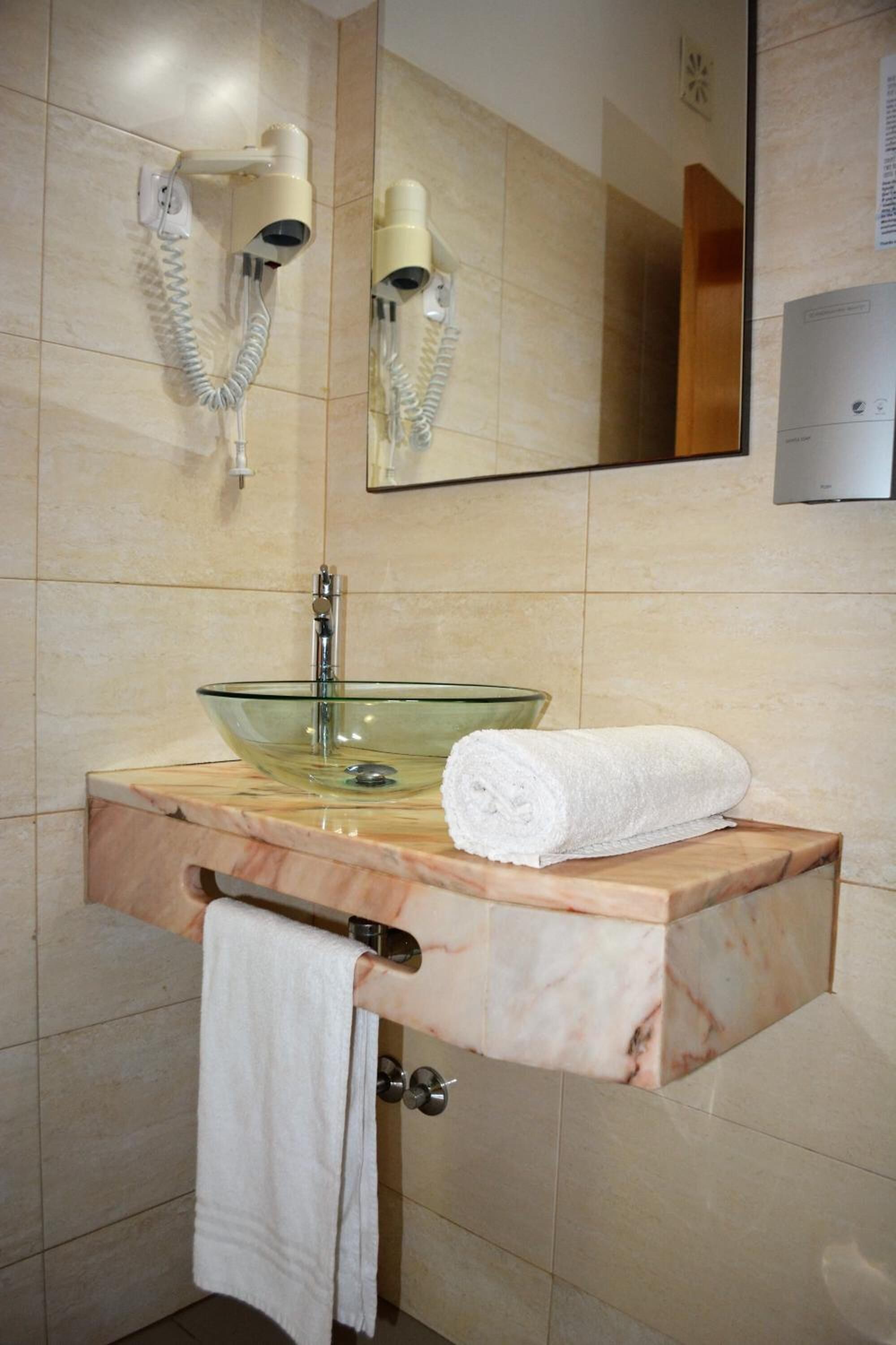 A bathroom with a glass bowl sink, a towel rack, and a mirror with wall-mounted hair dryers.