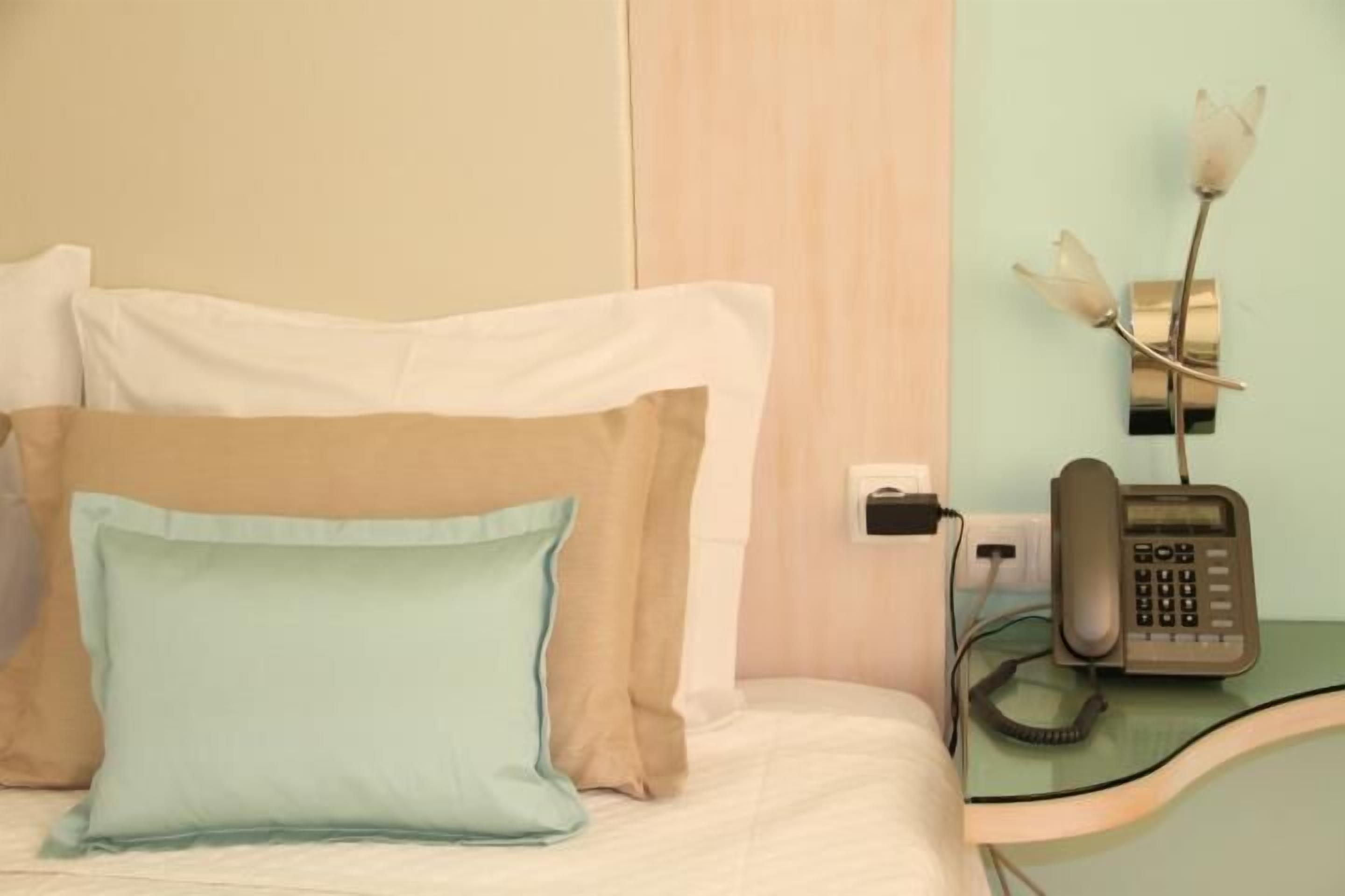 Hotellihuone, jossa on sänky, tyynyt, yöpöytä, jossa on puhelin, ja seinään kiinnitetty valaisin.