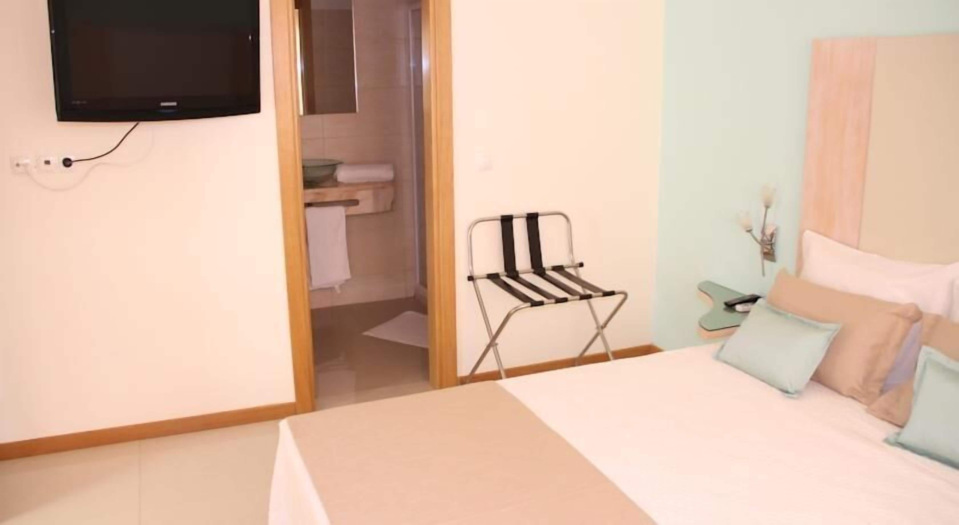 Un dormitorio con una cama, un televisor montado en la pared, una plancha pequeña y un baño visible a través de una puerta abierta.