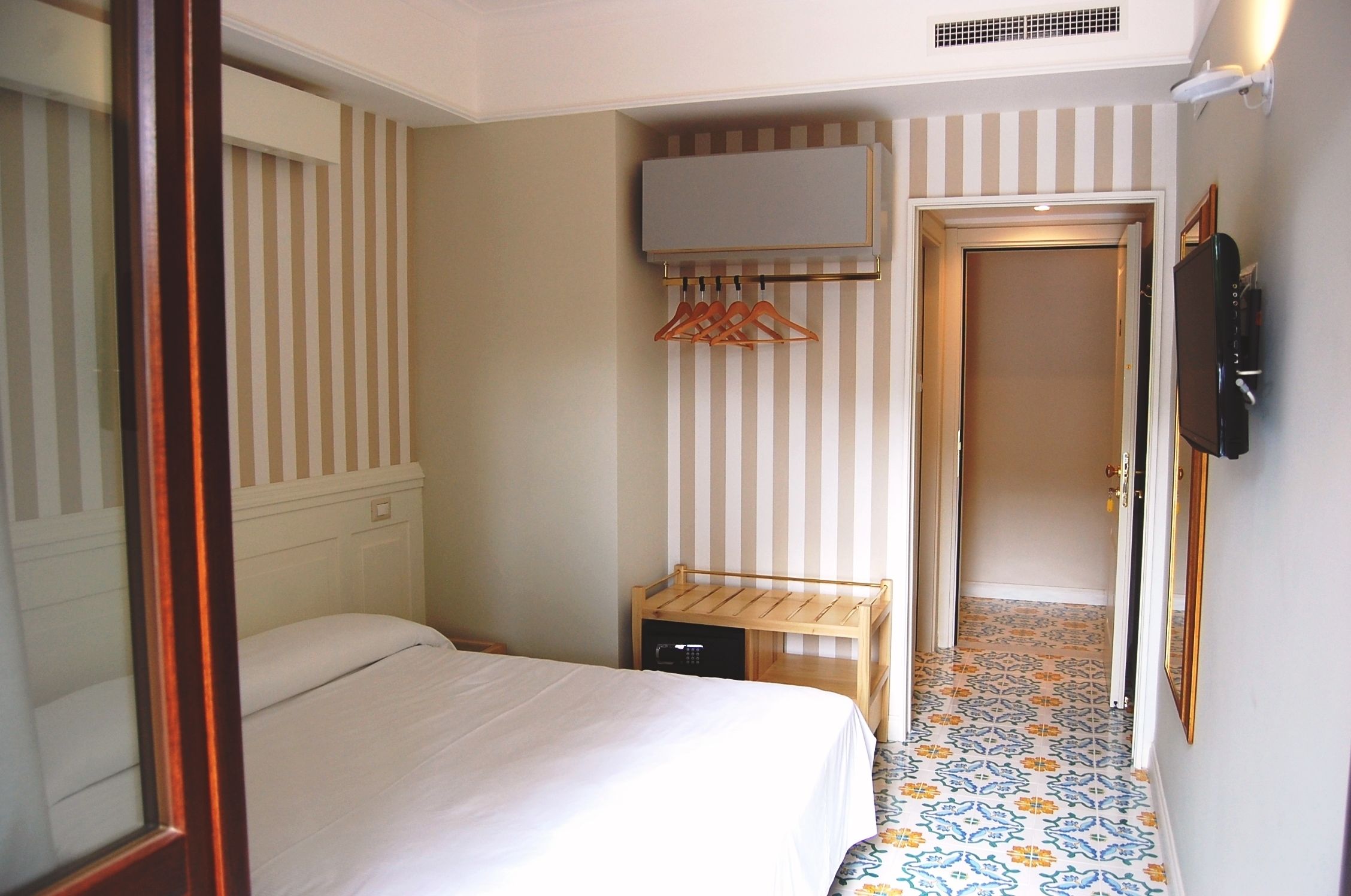 Una habitación de hotel con una cama, un televisor montado en la pared, un banco de madera y una puerta que da a otra habitación.