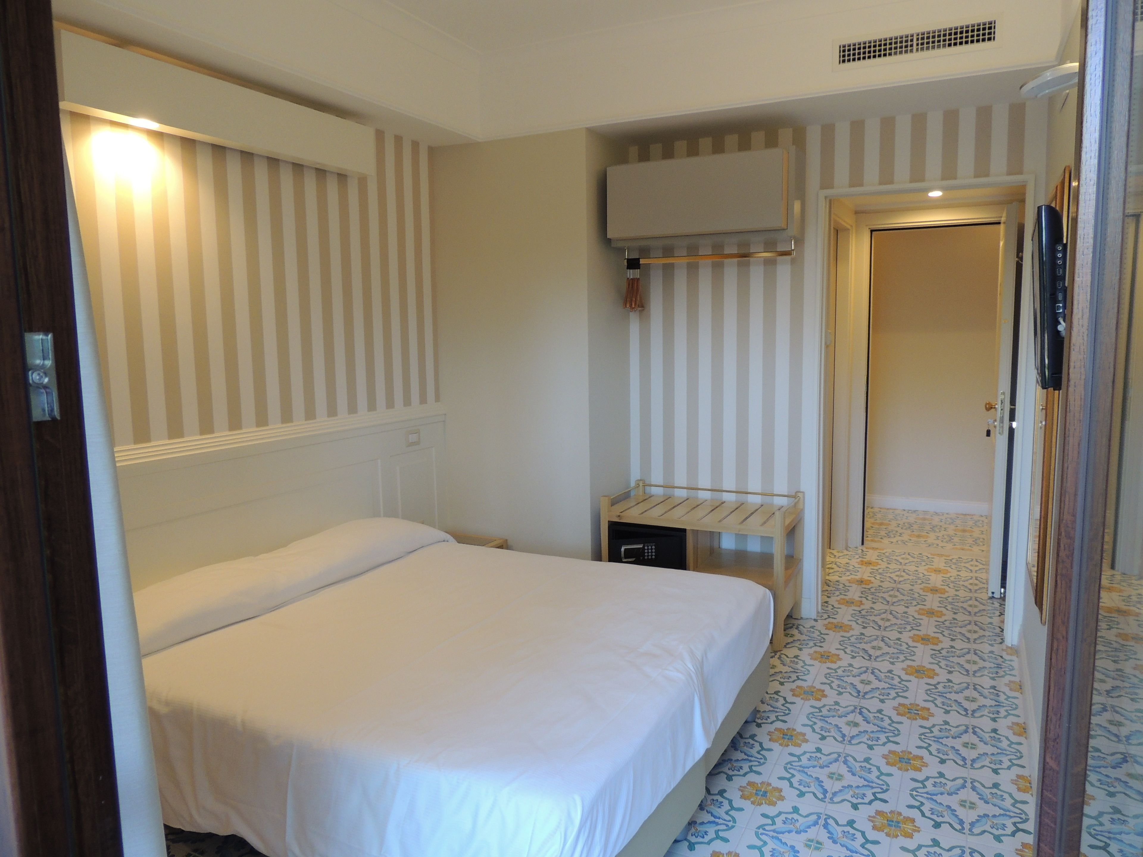 Quarto de hotel com uma cama, mesa de cabeceira, televisão na parede e porta de acesso a outro espaço.