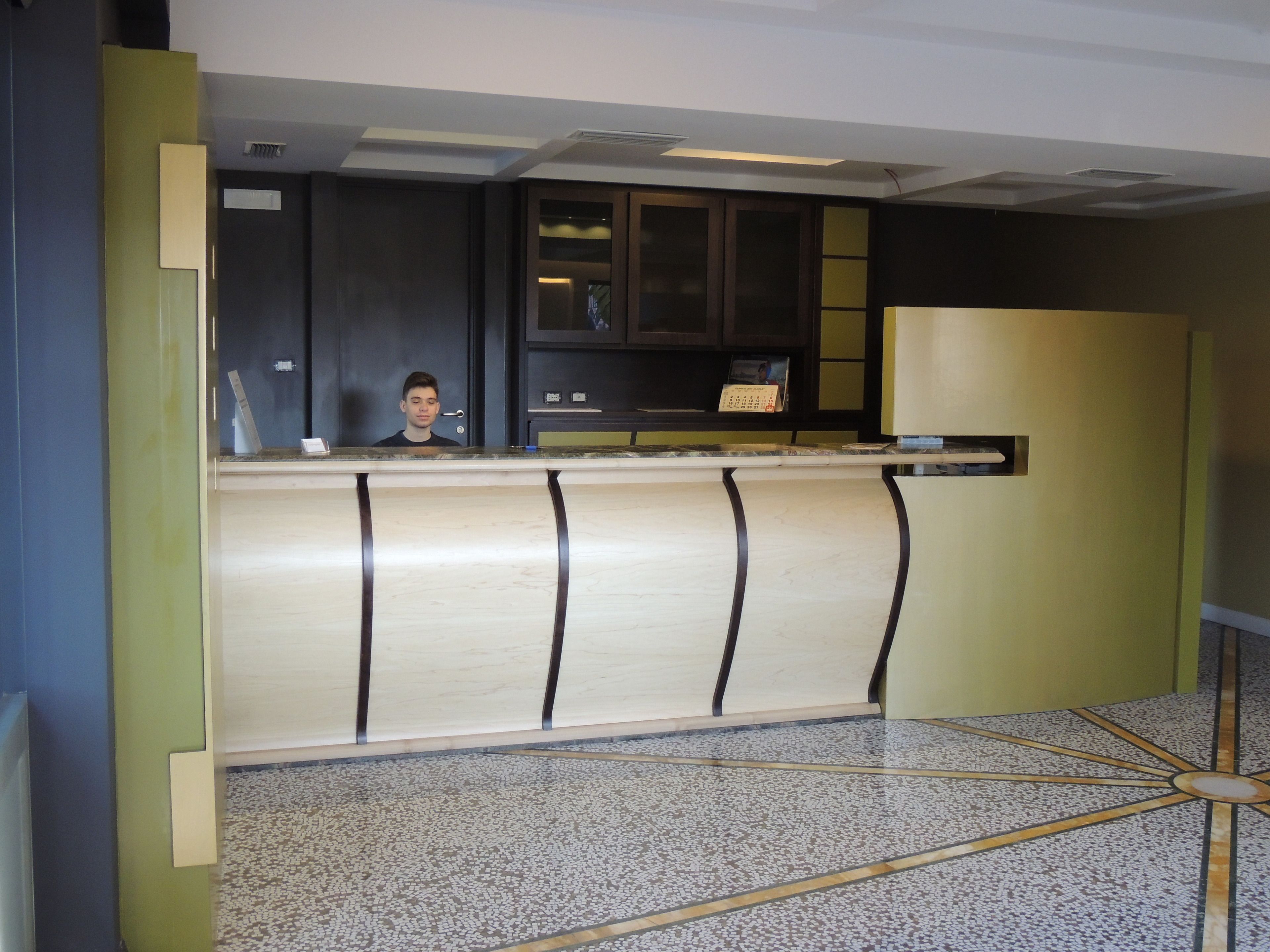 A reception desk with a person standing behind it, a dark wooden cabinet above, and a patterned floor.