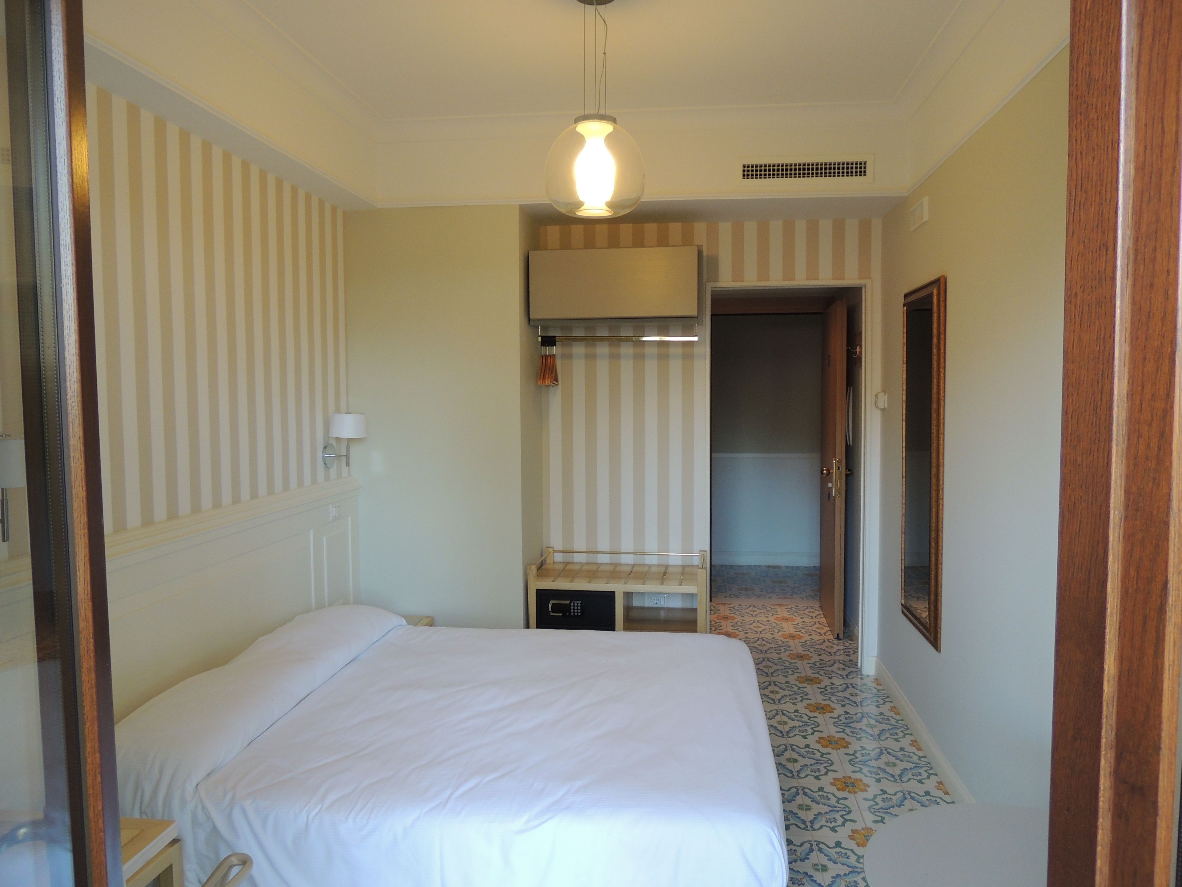 A hotel room with a bed, a small table, a lamp, a wardrobe, and a door leading to another room.