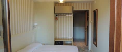 A hotel room with a bed, a small table, a lamp, a wardrobe, and a door leading to another room.