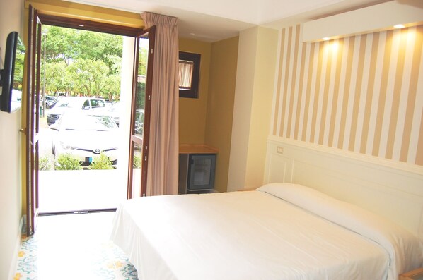 A hotel room with a bed, a television, and a view of parked cars and greenery outside. - Hotel Zeus Pompei (Pompei)