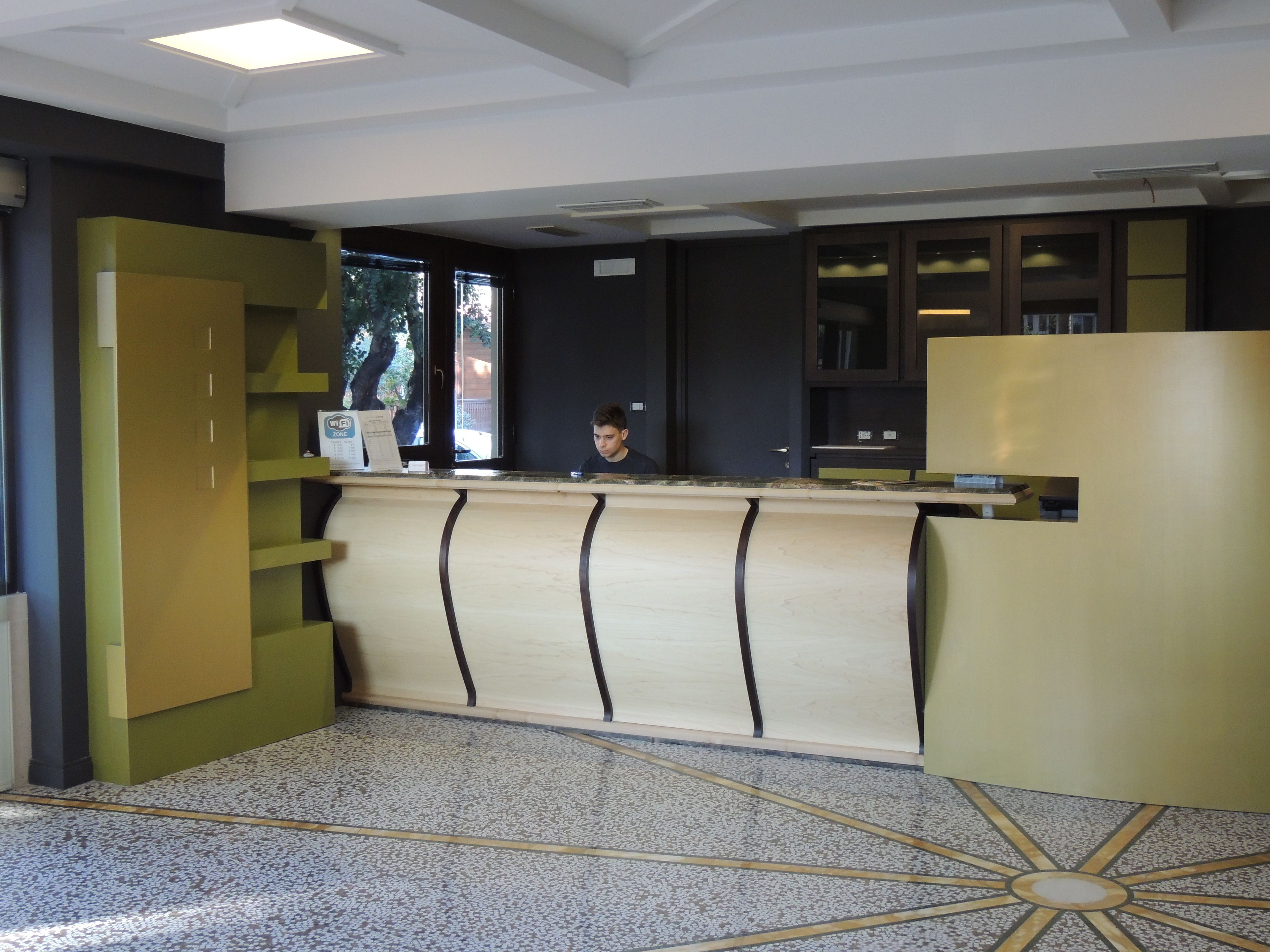 A reception desk with a modern design, a counter, and a computer. There are shelves and cabinets to the left, and a large window to the right.
