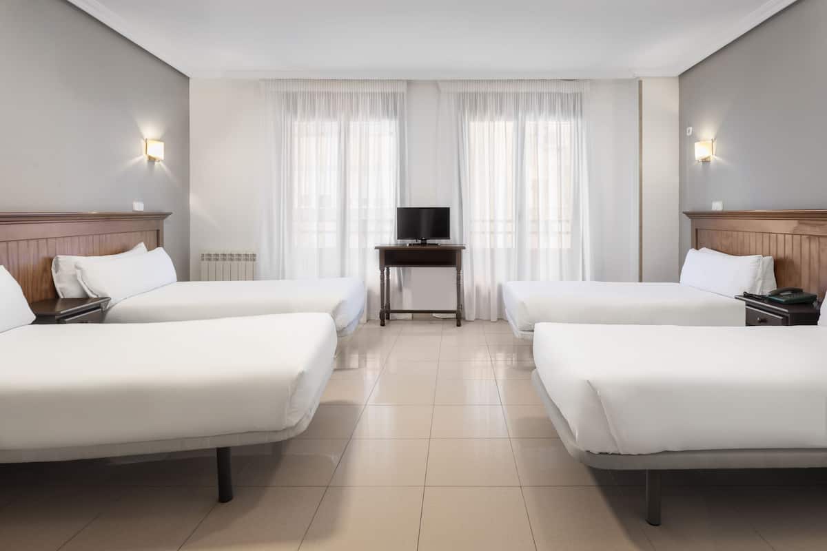 Una habitación de hotel con dos camas individuales, una mesita con un televisor y una ventana con cortinas de gasa.