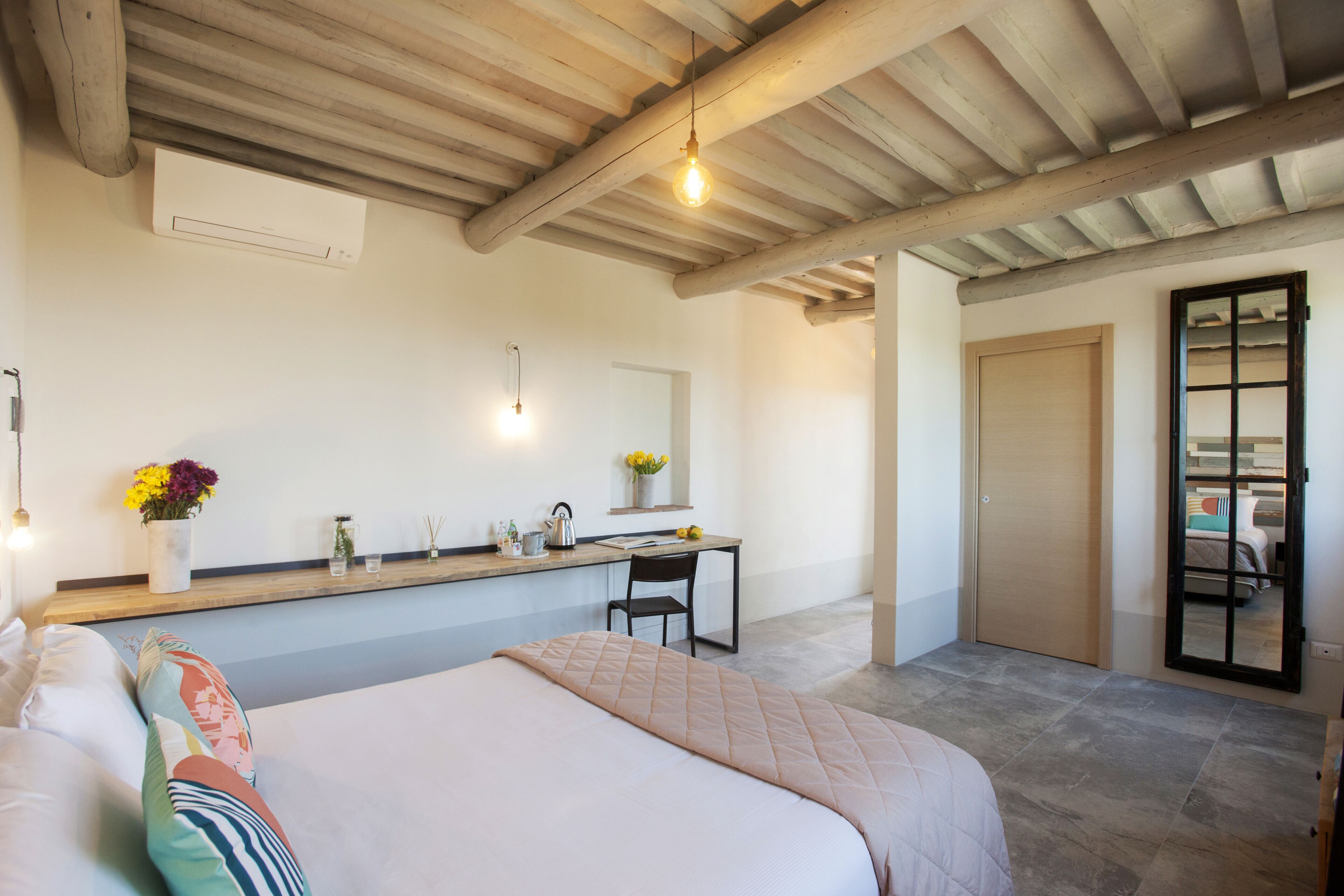 Quarto com cama, escrivaninha e cadeira. Há uma janela, uma porta e um teto com vigas de madeira.