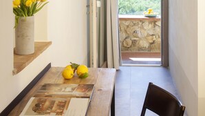 A narrow hallway with a wooden table, a chair, a vase with yellow flowers, and a magazine. A door leads to a balcony with a stone wall and a view of greenery. - Casale Sterpeti (Magliano in Toscana)