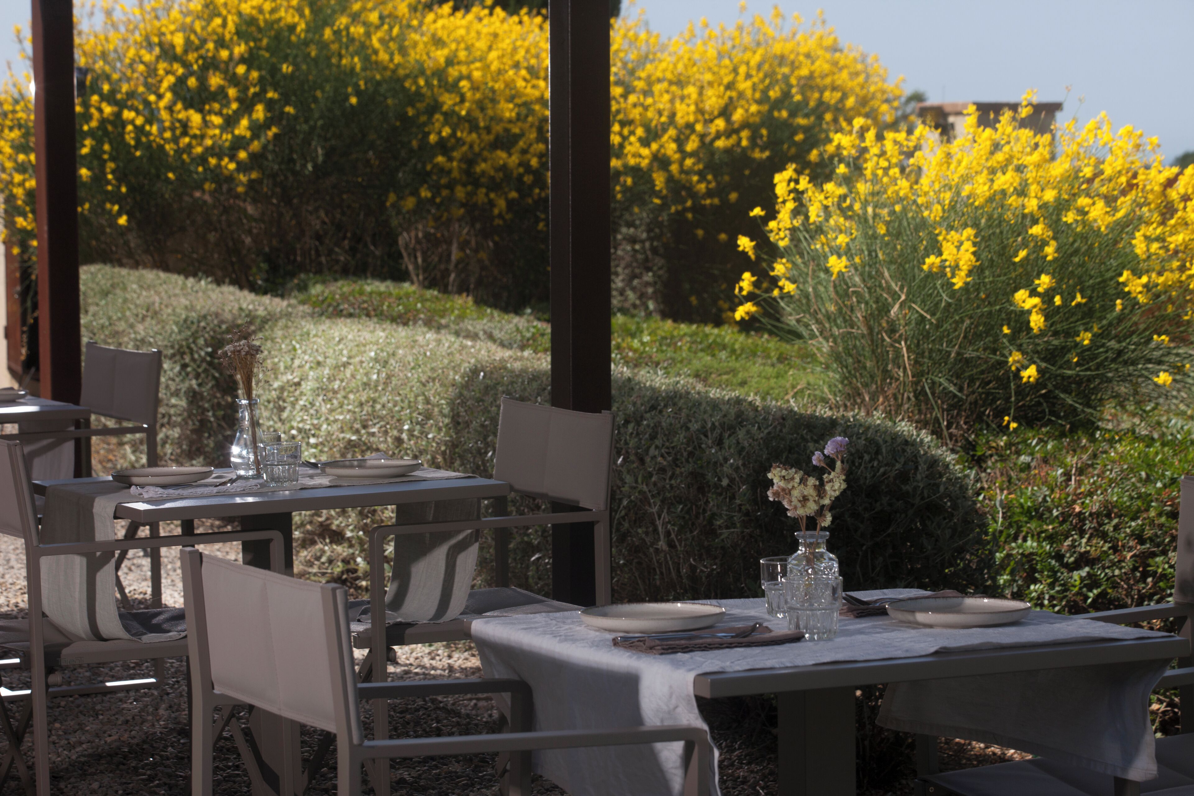 An outdoor dining area with tables set for two, surrounded by a garden with yellow flowers.