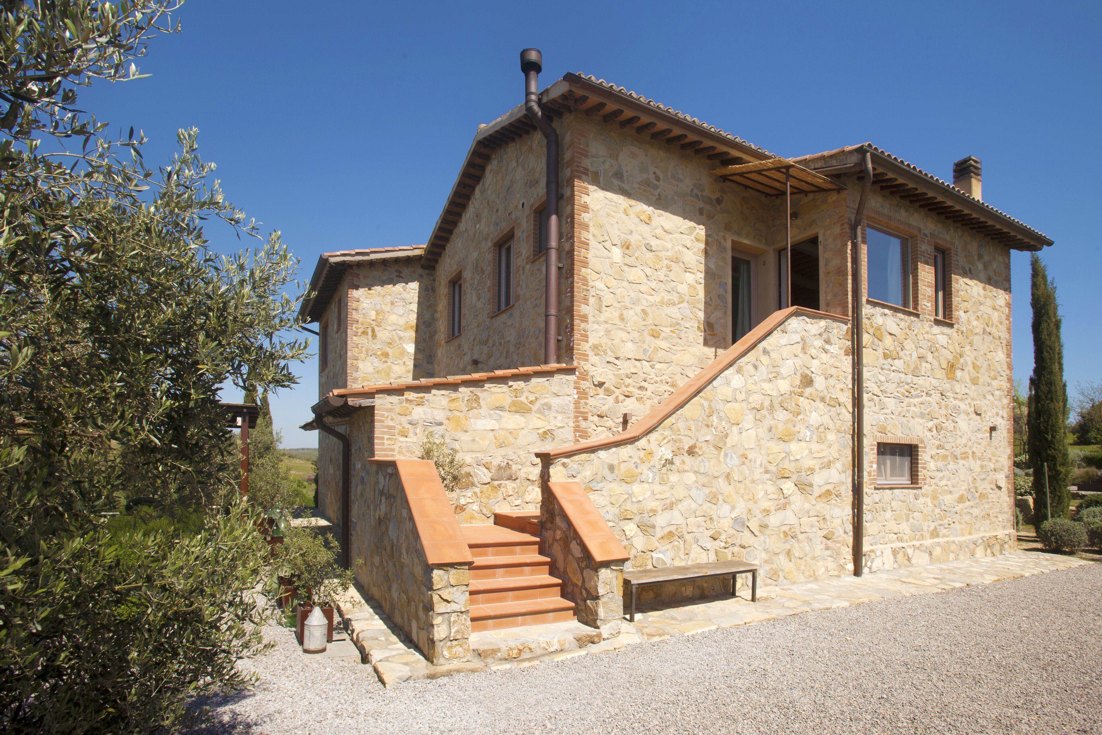 Un edificio in pietra con una scalinata che conduce all'ingresso, circondato da ulivi e un cielo sereno.