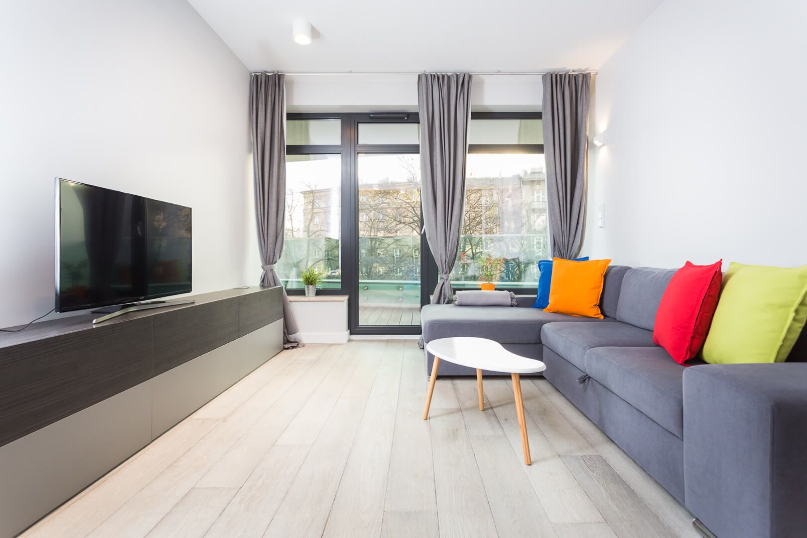 A modern living room with a flat-screen TV, grey sofa, colorful cushions, and a small white table.