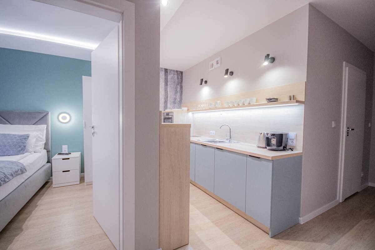 A modern bedroom with a bed, bedside table, and a nightstand. A kitchenette with a sink and a coffee machine is visible.