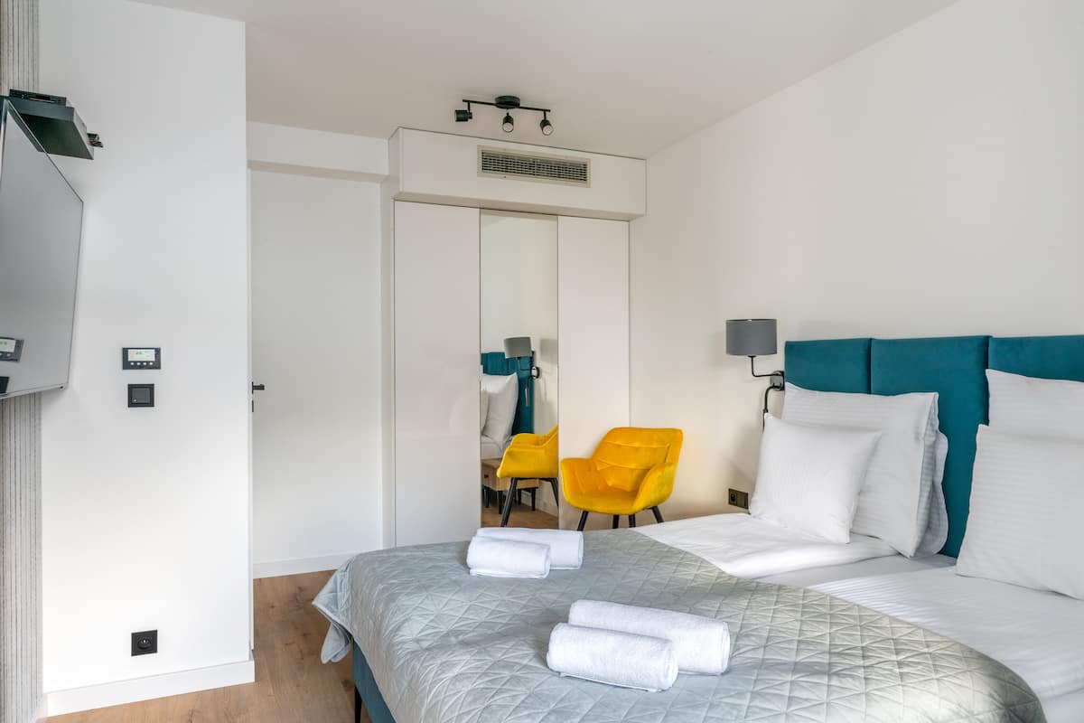 A hotel room with a bed, a chair, a mirror, and a wall-mounted control panel.