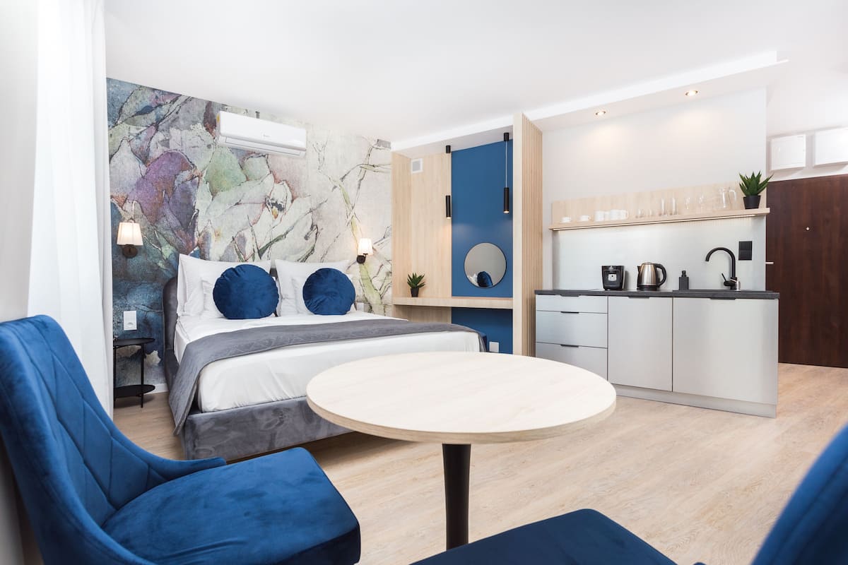 A modern hotel room with a bed, a round table, and a kitchenette.