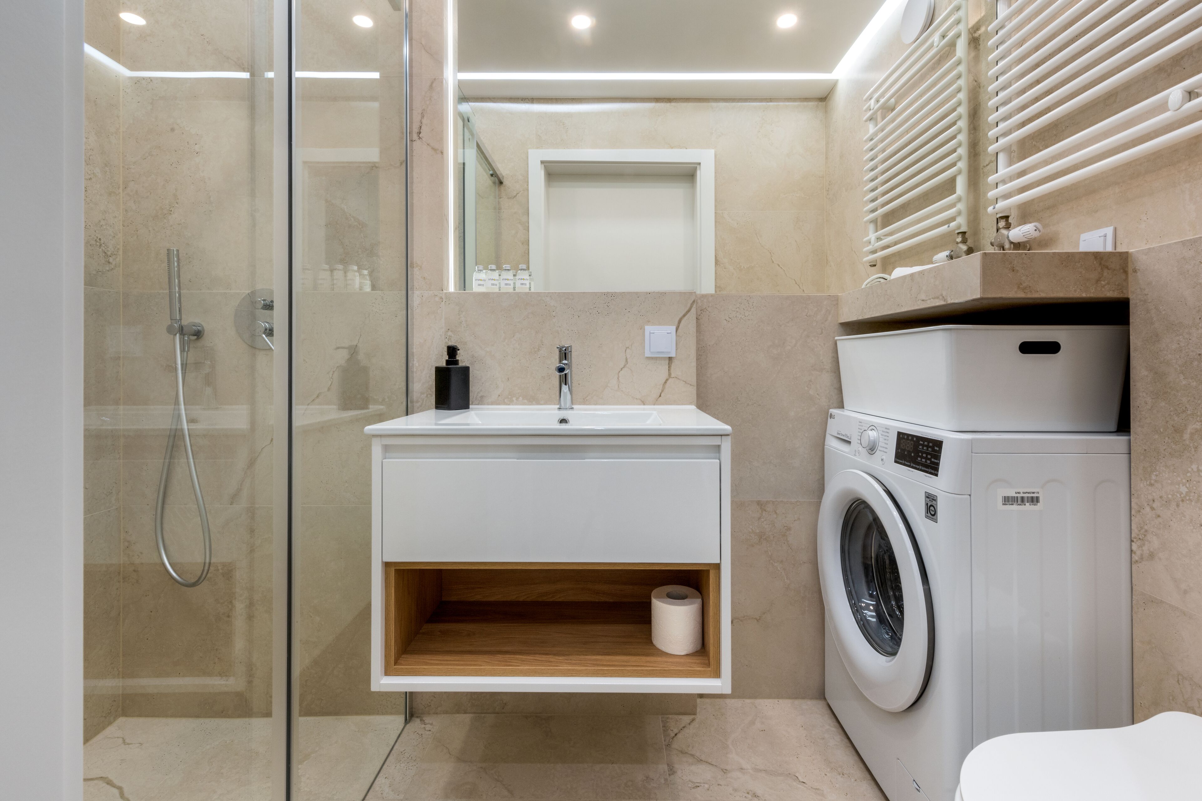 A modern bathroom with a glass-enclosed shower, a built-in sink with a wooden shelf, and a stacked washer and dryer.