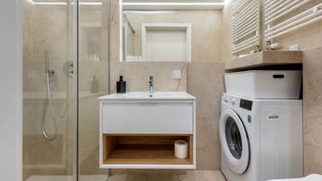 A modern bathroom with a glass-enclosed shower, a built-in sink with a wooden shelf, and a stacked washer and dryer.