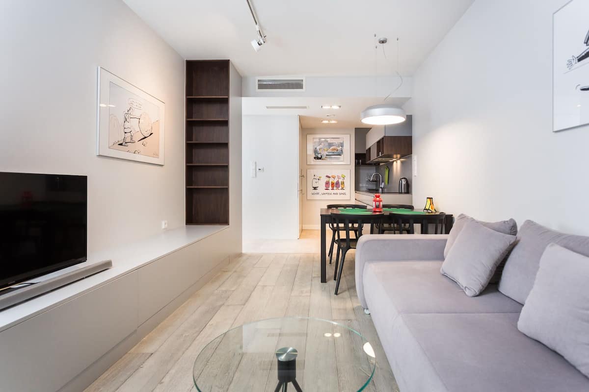 A modern living room with a flat-screen TV, a glass coffee table, a grey sofa, and a dining area with a table and chairs.