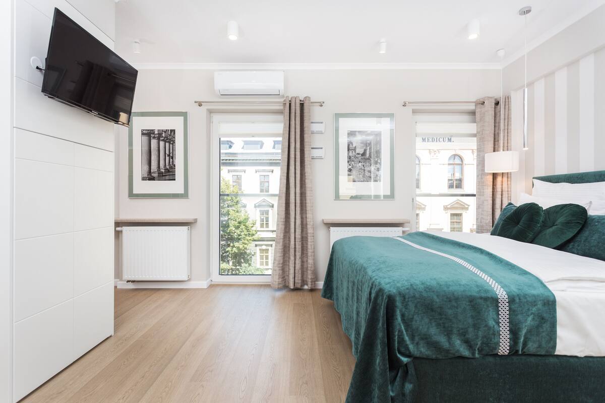 A modern bedroom with a bed, a TV mounted on the wall, and a window with a view of buildings outside.