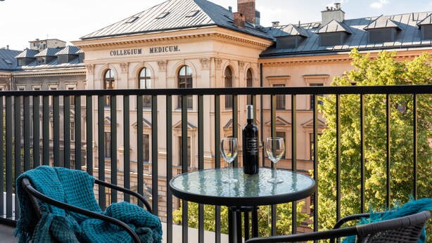 Un balcón con una mesa y sillas, una botella de vino, y un edificio con la inscripción 'COLLEGIUM MEDICUM' al fondo.