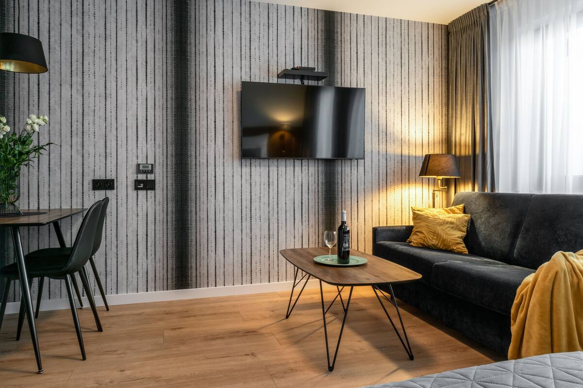 A modern living room with a flat-screen TV mounted on a striped wallpaper wall, a wooden coffee table, a black sofa with yellow cushions, and a lamp.