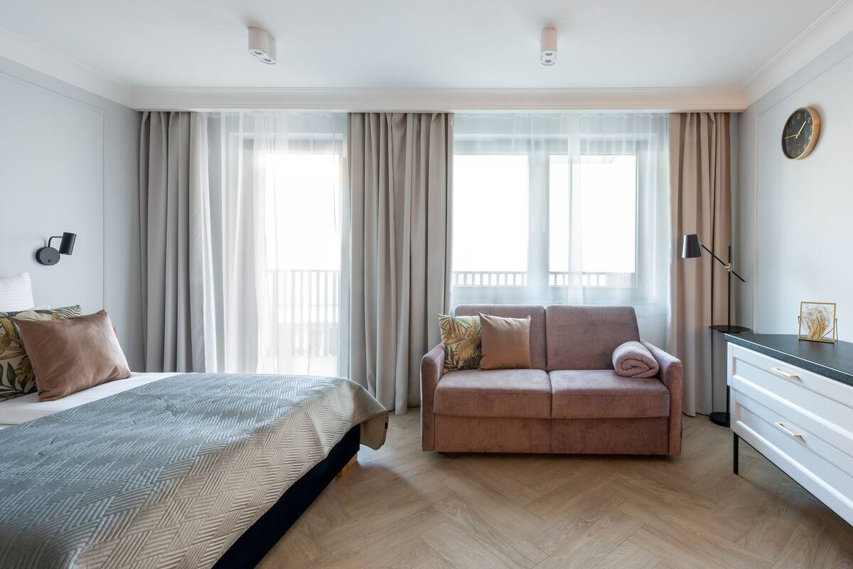 A modern hotel room with a bed, a sofa, a desk, and a large window with curtains.