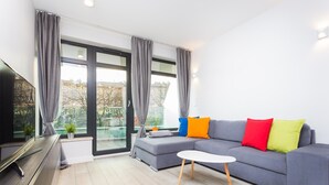 Una sala de estar moderna con un sofá gris, cojines coloridos, una mesa de centro blanca y un gran ventanal con cortinas.