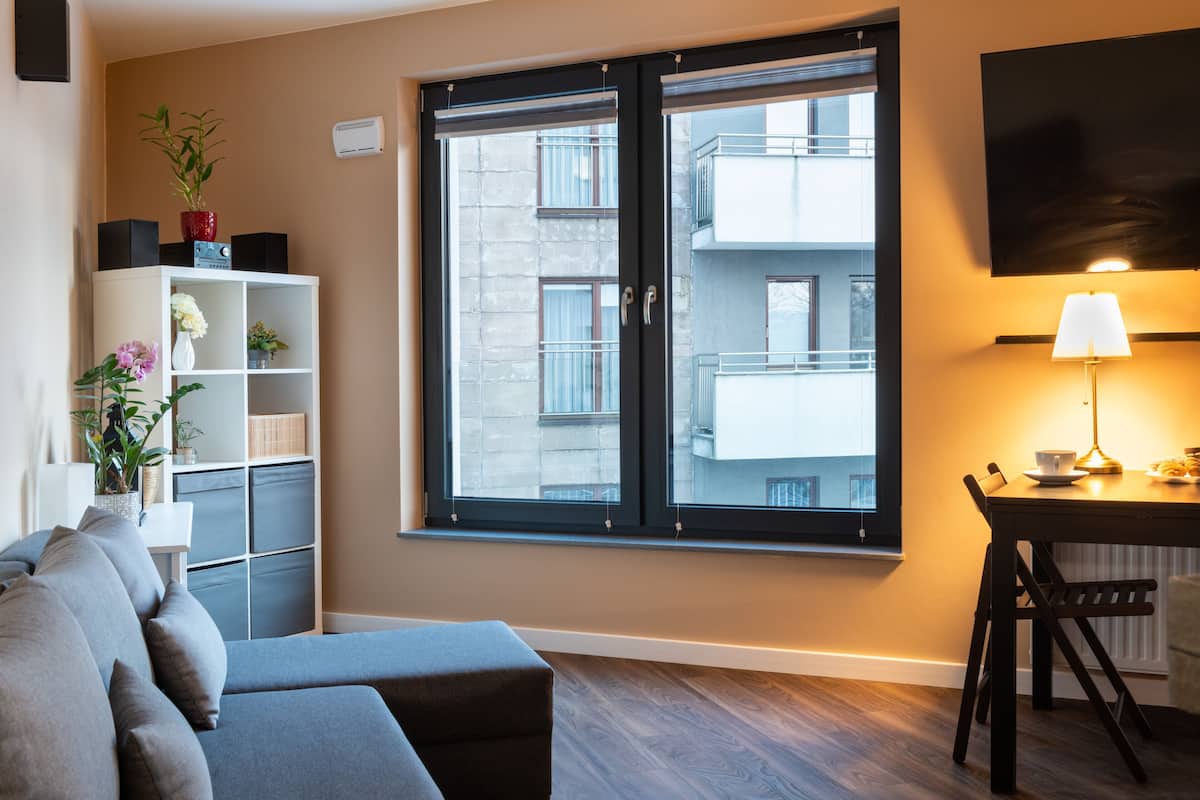 A modern living room with a grey sofa, a white shelving unit with plants, and a large window with a view of buildings.