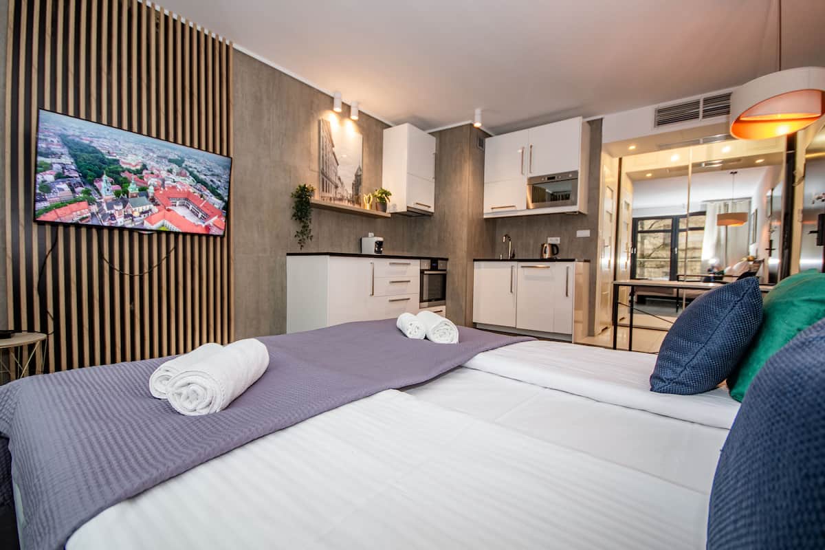 A hotel room with a bed, a TV mounted on the wall, a kitchenette, and a bathroom visible through an open door.
