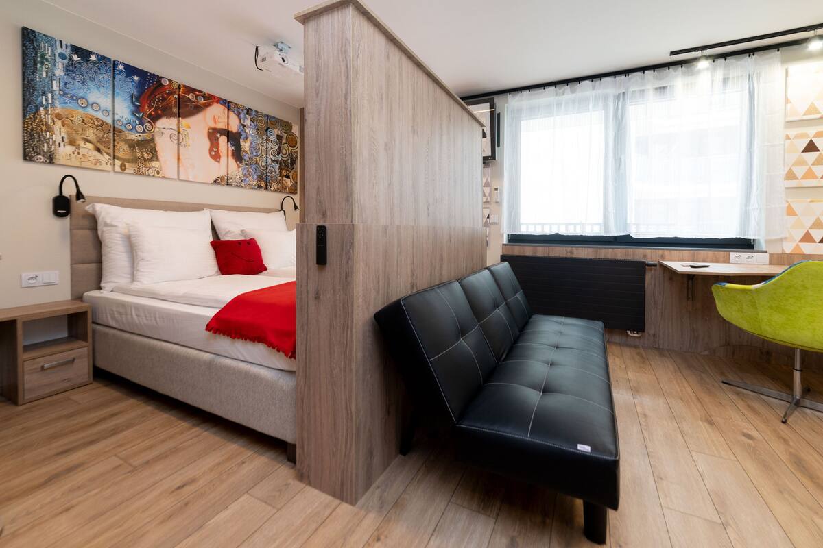 A compact room with a bed, a black sofa, a green chair, and a painting on the wall.