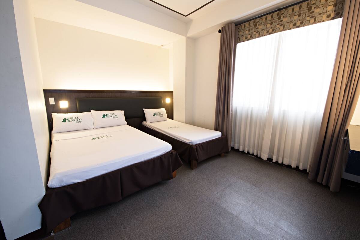 Una habitación de hotel con dos camas individuales, cabeceras, una ventana con cortinas y suelo alfombrado.