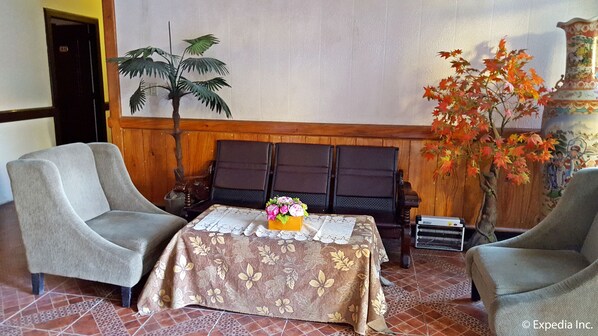 Une pièce comprenant deux fauteuils gris, un banc en bois, une table recouverte d’une nappe à motifs floraux, une plante en pot et un vase décoratif.