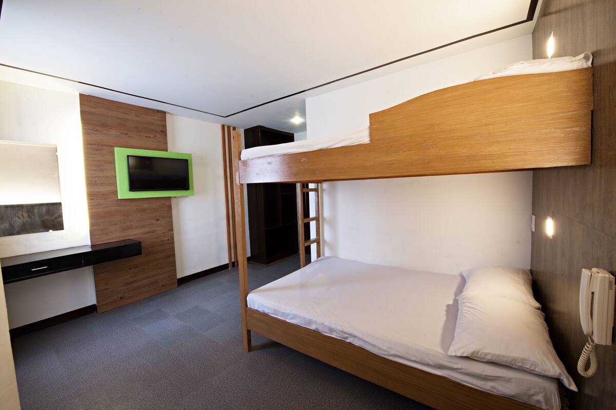 Habitación con literas, cabecero de madera, teléfono en la mesita de noche y televisor con marco verde en la pared.
