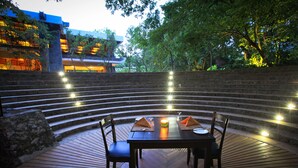 Un área de comedor al aire libre circular con una mesa y sillas, rodeada por un muro de piedra y escalones que conducen a un edificio.