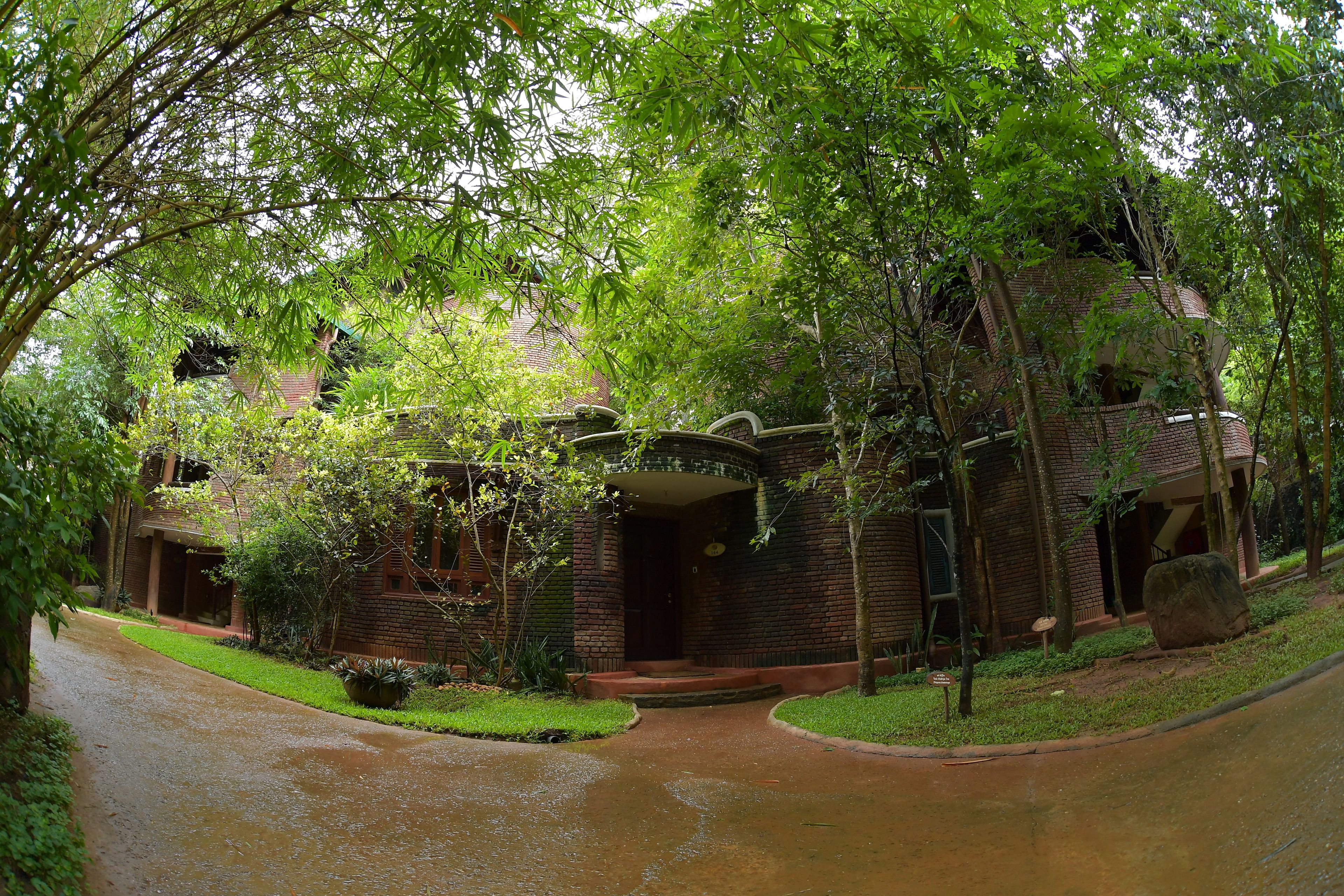 A traditional brick house with a curved entrance, surrounded by lush greenery and a wet driveway.