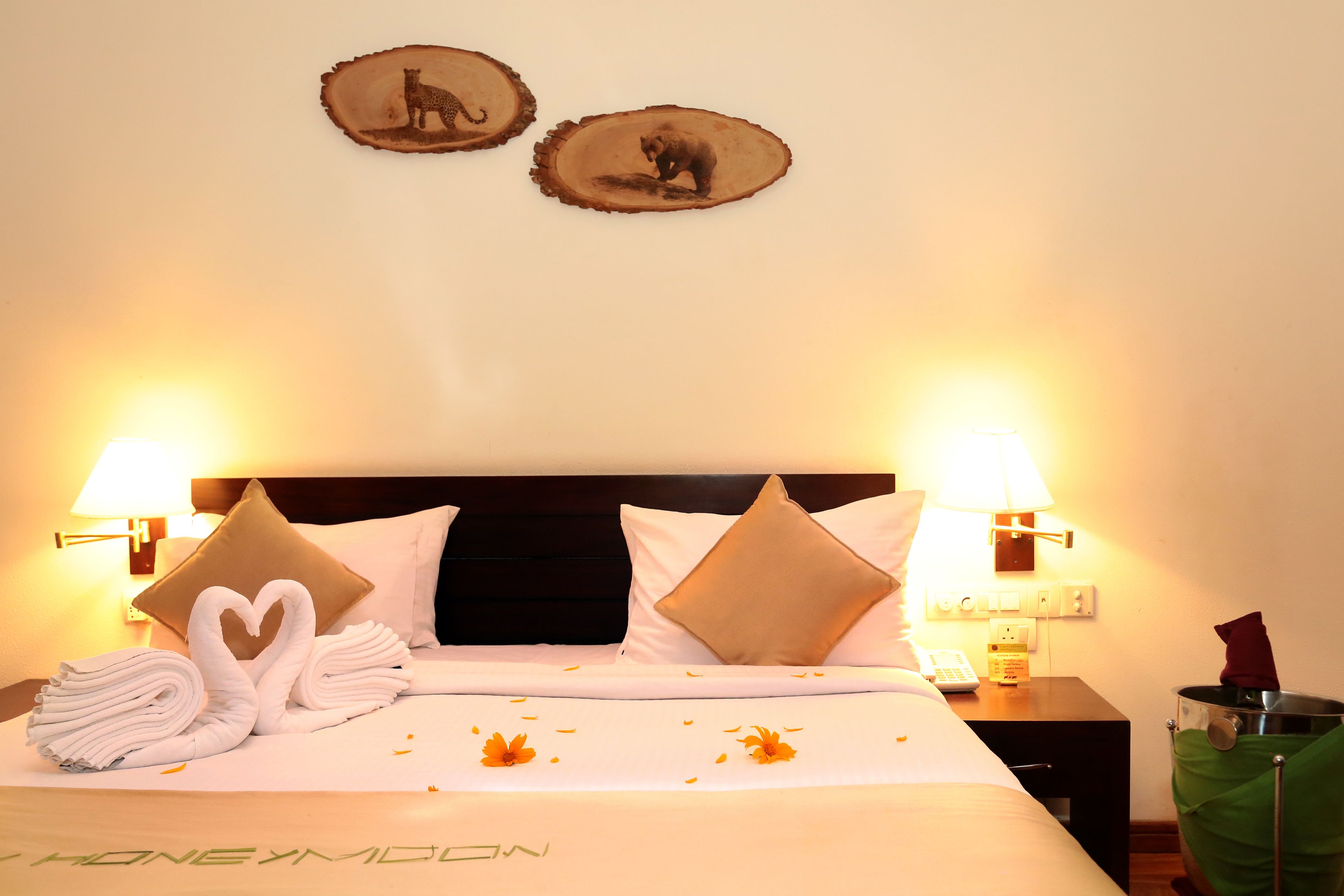 A neatly made bed with white linens, two heart-shaped towels, and a bedspread with orange flowers. There are two wall-mounted lamps, a nightstand with a phone, and a green bag hanging on the wall.