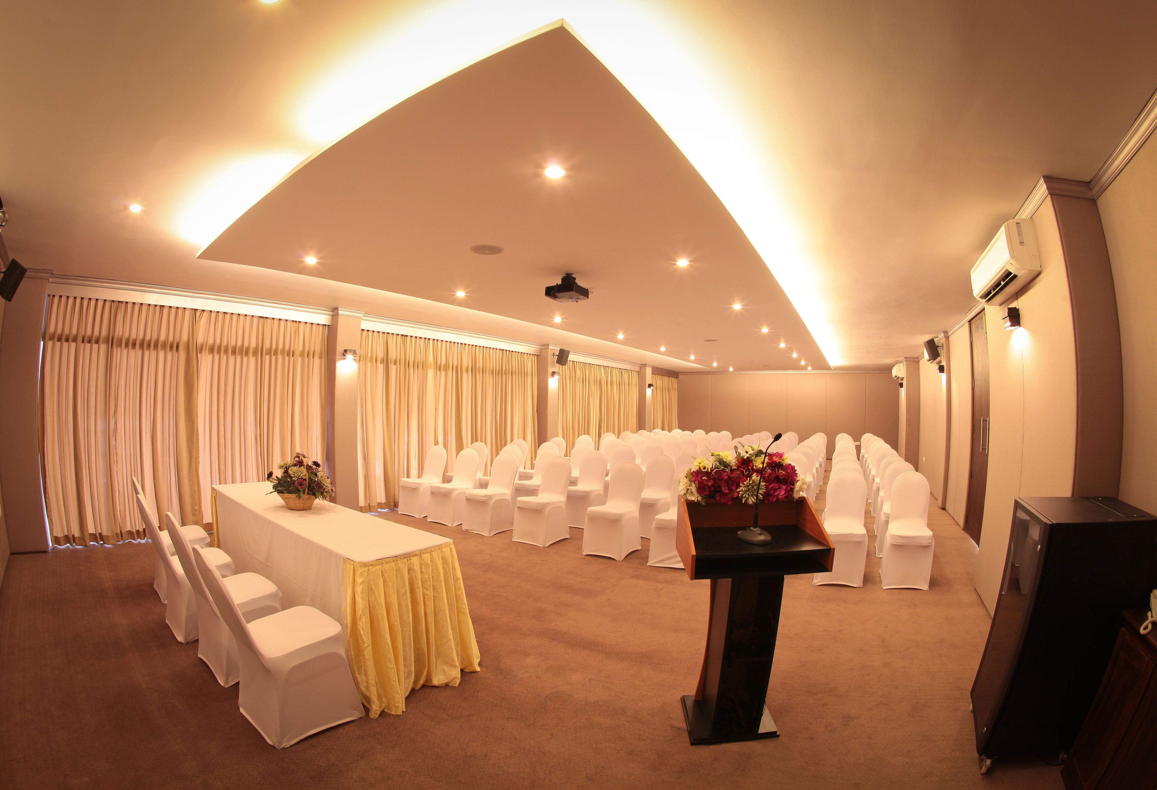 A conference room with rows of white chairs, a long table with a yellow tablecloth, and a podium with flowers.